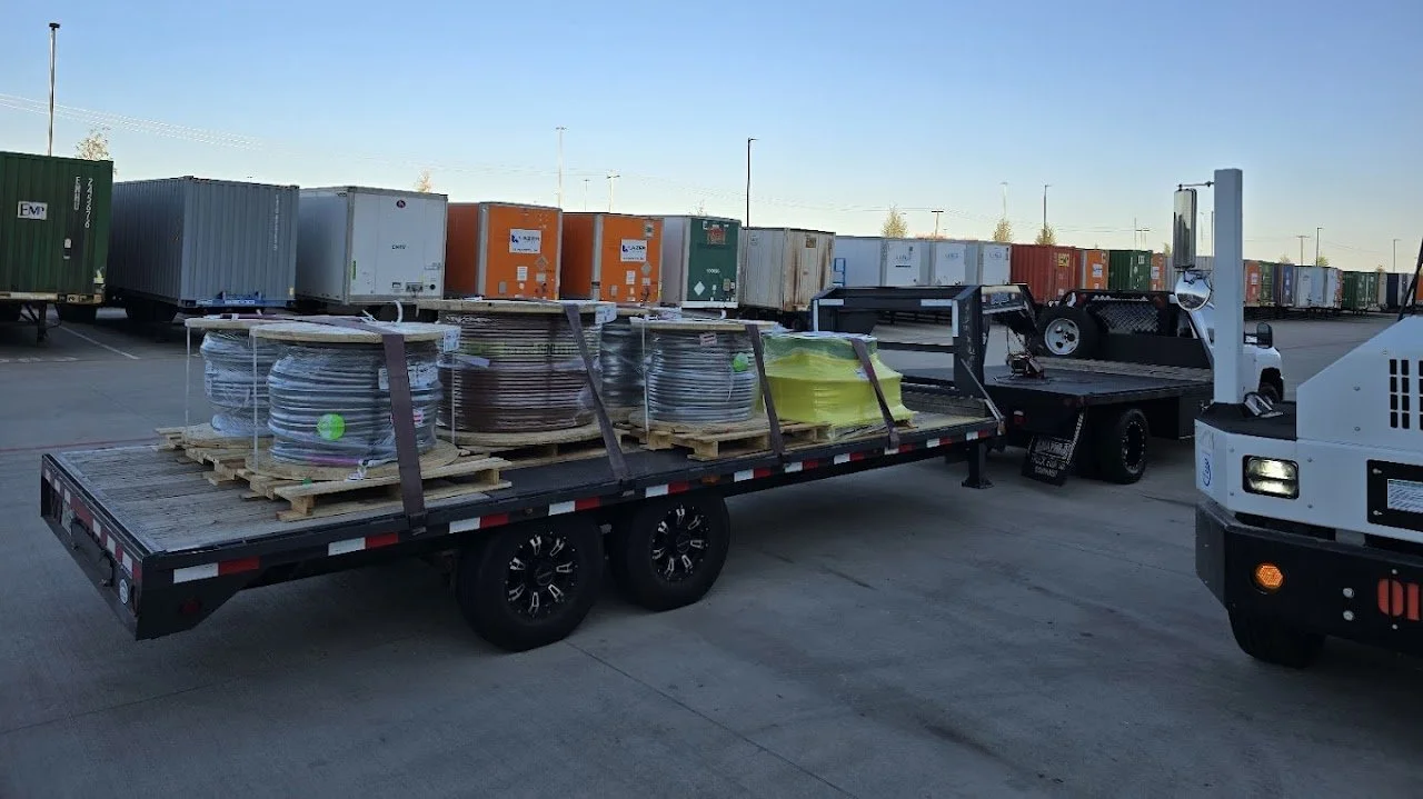 Flatbed truck carrying large coils of wiring and yellow hoses parked in a lot with shipping containers in the background.