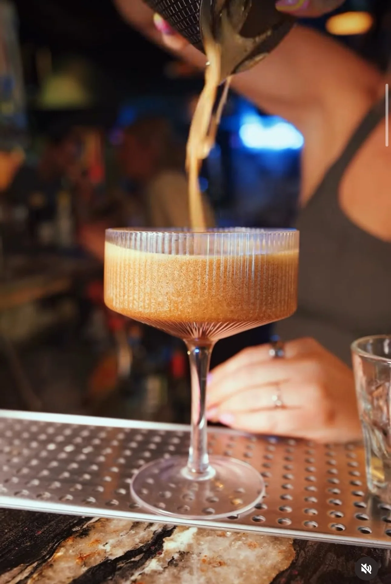 A bartender pouring a foamy, espresso martini into a coupe glass at a bar, with a blurred background of other bar patrons.