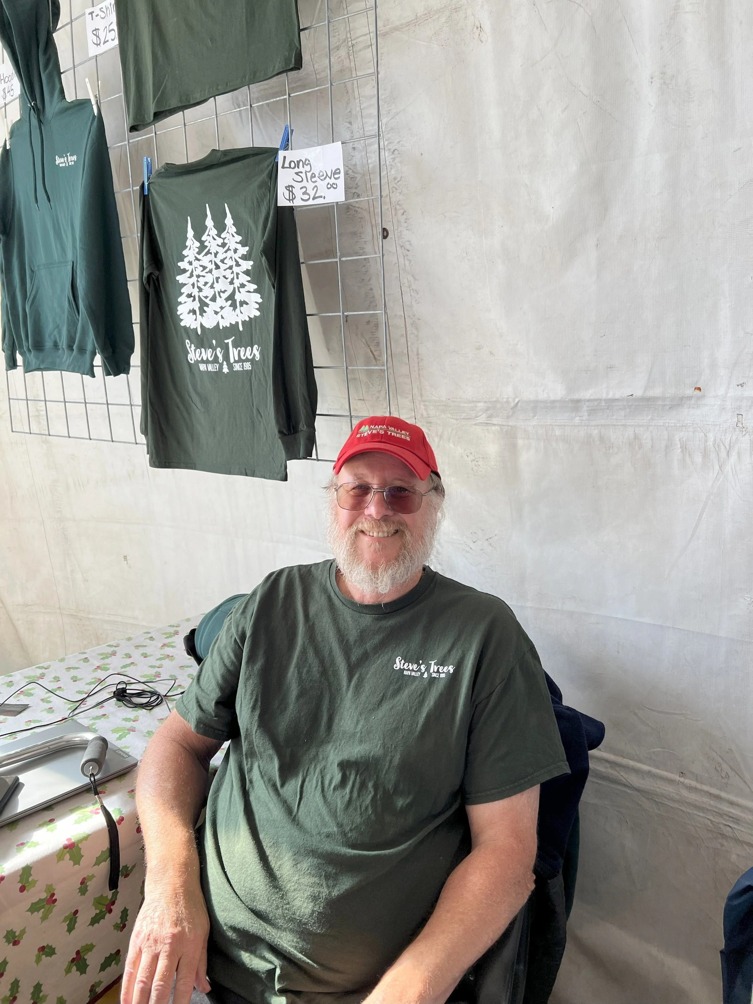 A smiling man with a beard wearing glasses, a red cap, and a green T-shirt sitting at a table inside a tent. On the table are a laptop, a microphone, and some wires. Behind him, a display with shirts and hoodies hanging on a wire grid, including a dark green hoodie and T-shirt with a white tree logo and the words 'Steve's Trees.' A small sign indicates the T-shirt costs $25, and a long sleeve shirt costs $32.