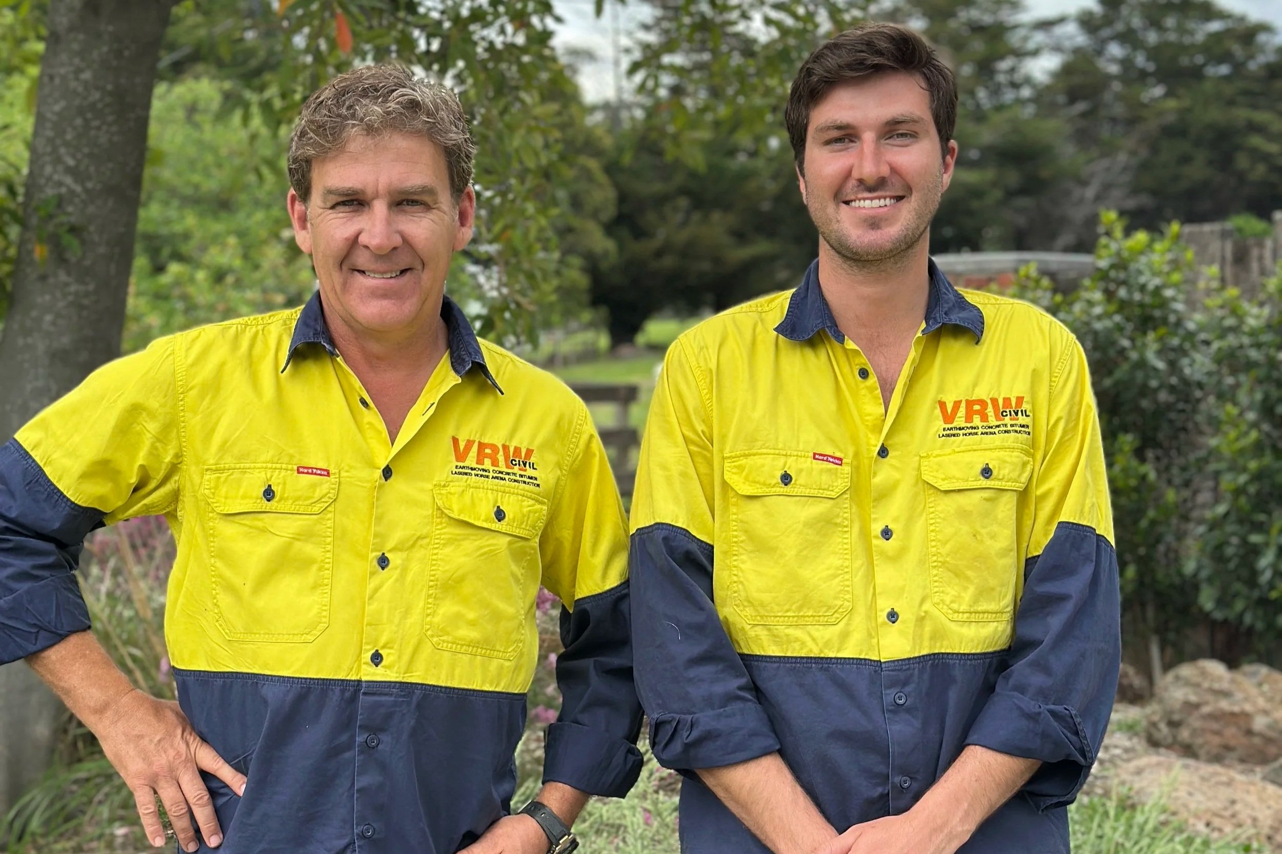 Two men in yellow and navy blue work shirts standing outdoors with trees and greenery in the background.