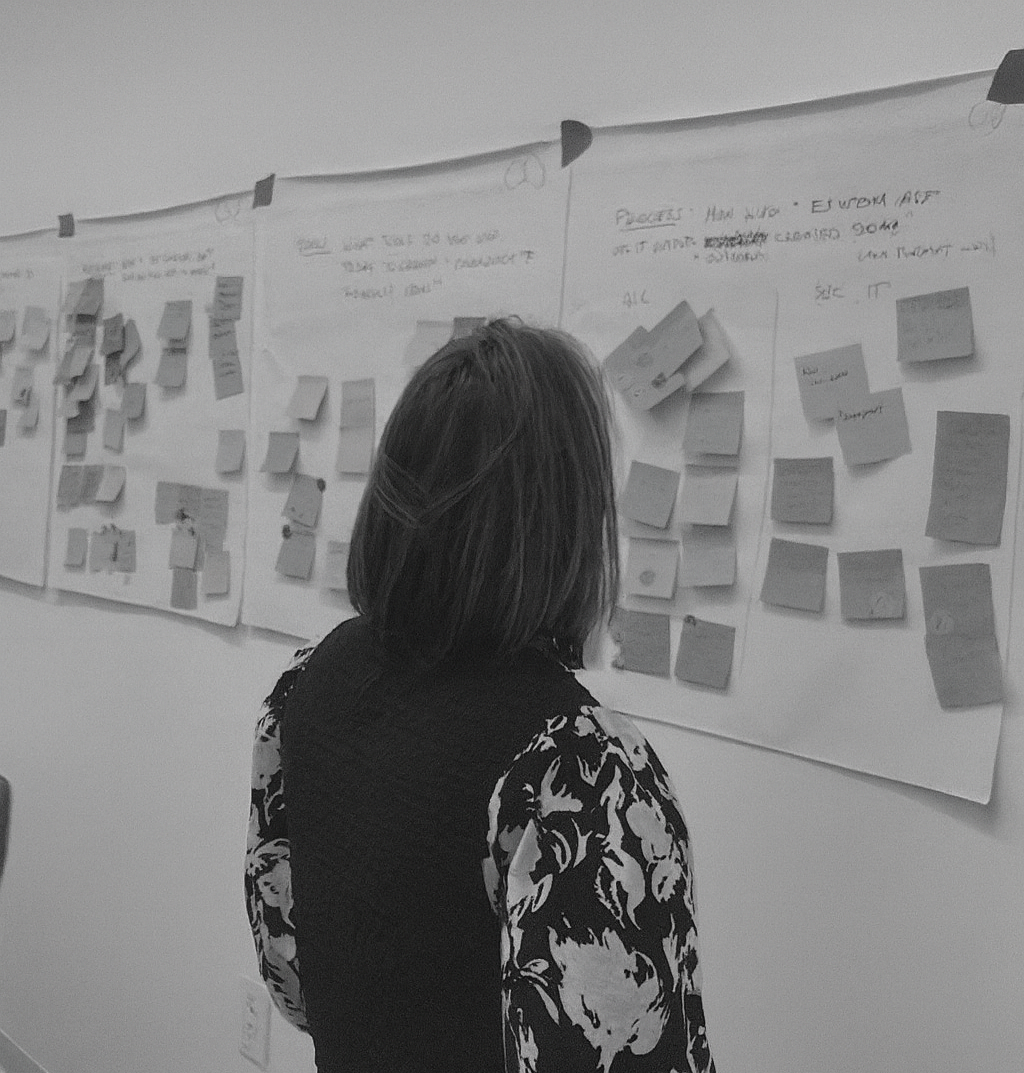 A woman with shoulder-length hair wearing a patterned top, standing in front of a wall covered with large sheets of paper filled with handwritten notes and numerous sticky notes, likely participating in a brainstorming or planning session.