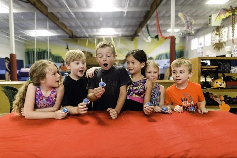 Five children gathered around a table with cupcakes, celebrating indoors, with colorful decorations overhead.