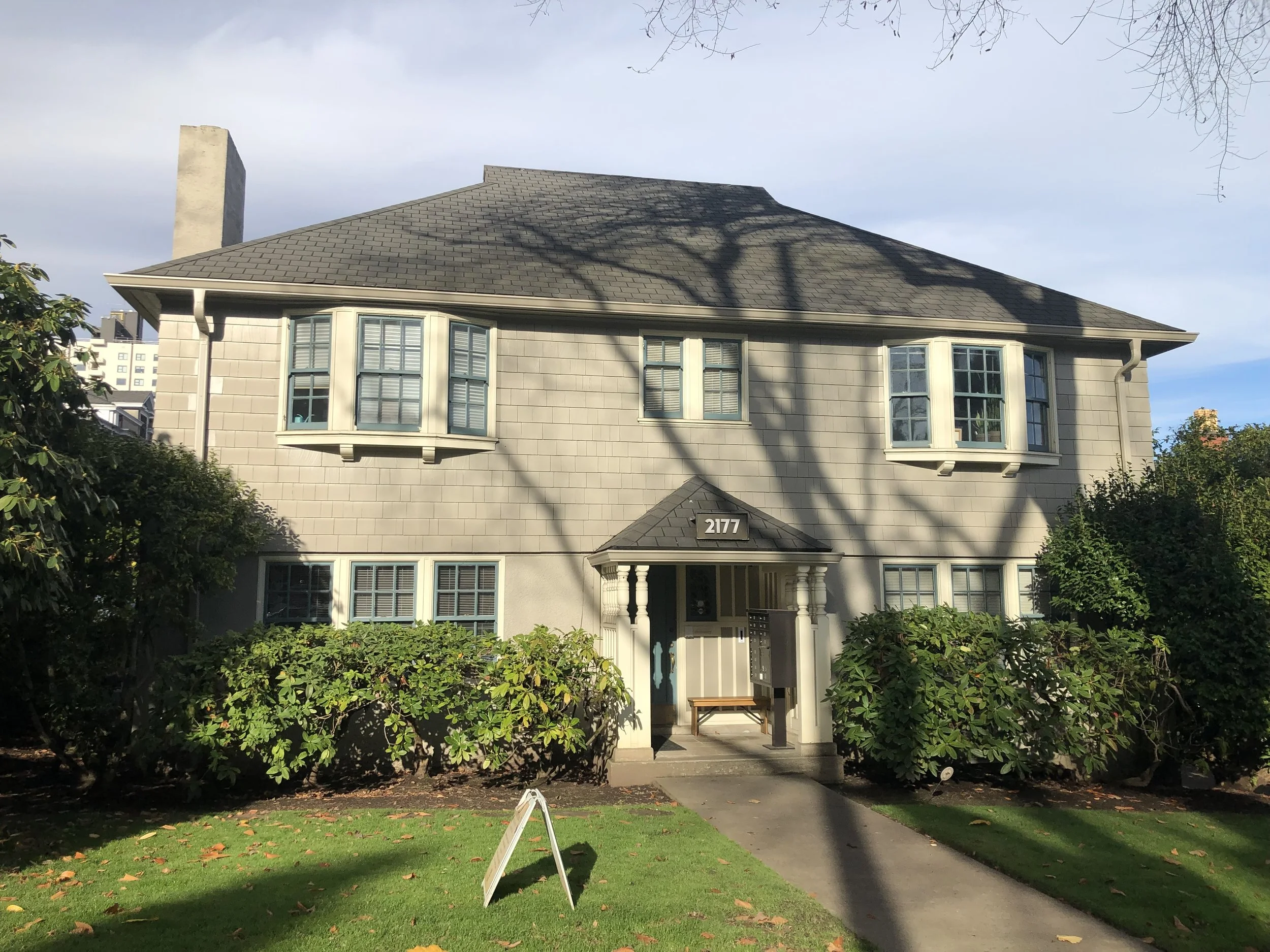 A two-story house with beige siding, blue-shuttered windows, a dark roof, and a front porch with a bench. There are bushes and a grassy lawn in front, and a sidewalk leading to the entrance. The house number '2177' is on the porch.