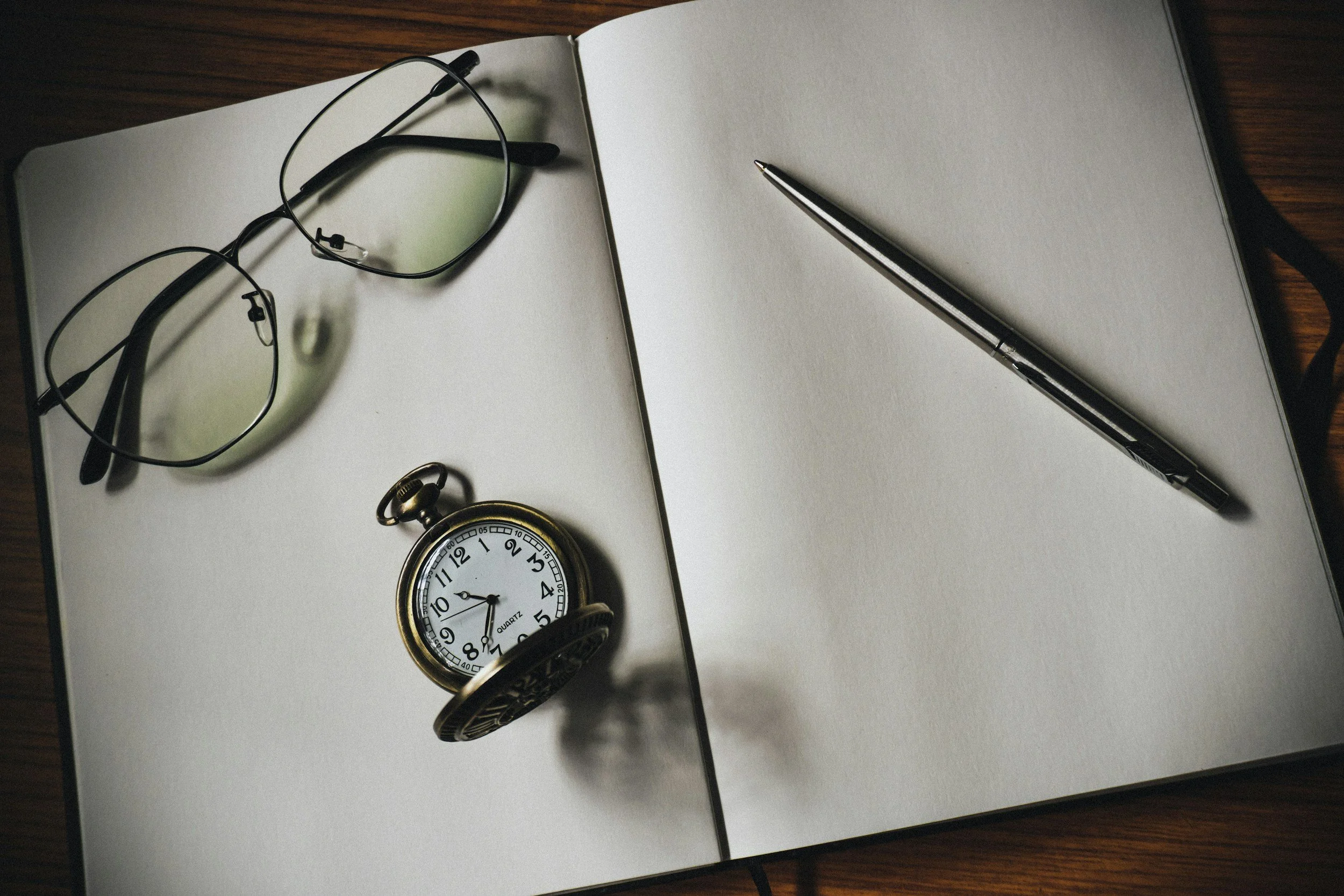 Open notebook with blank pages, used for freelance writing and editing, a pair of glasses, a pen, and a pocket watch on a wooden surface.