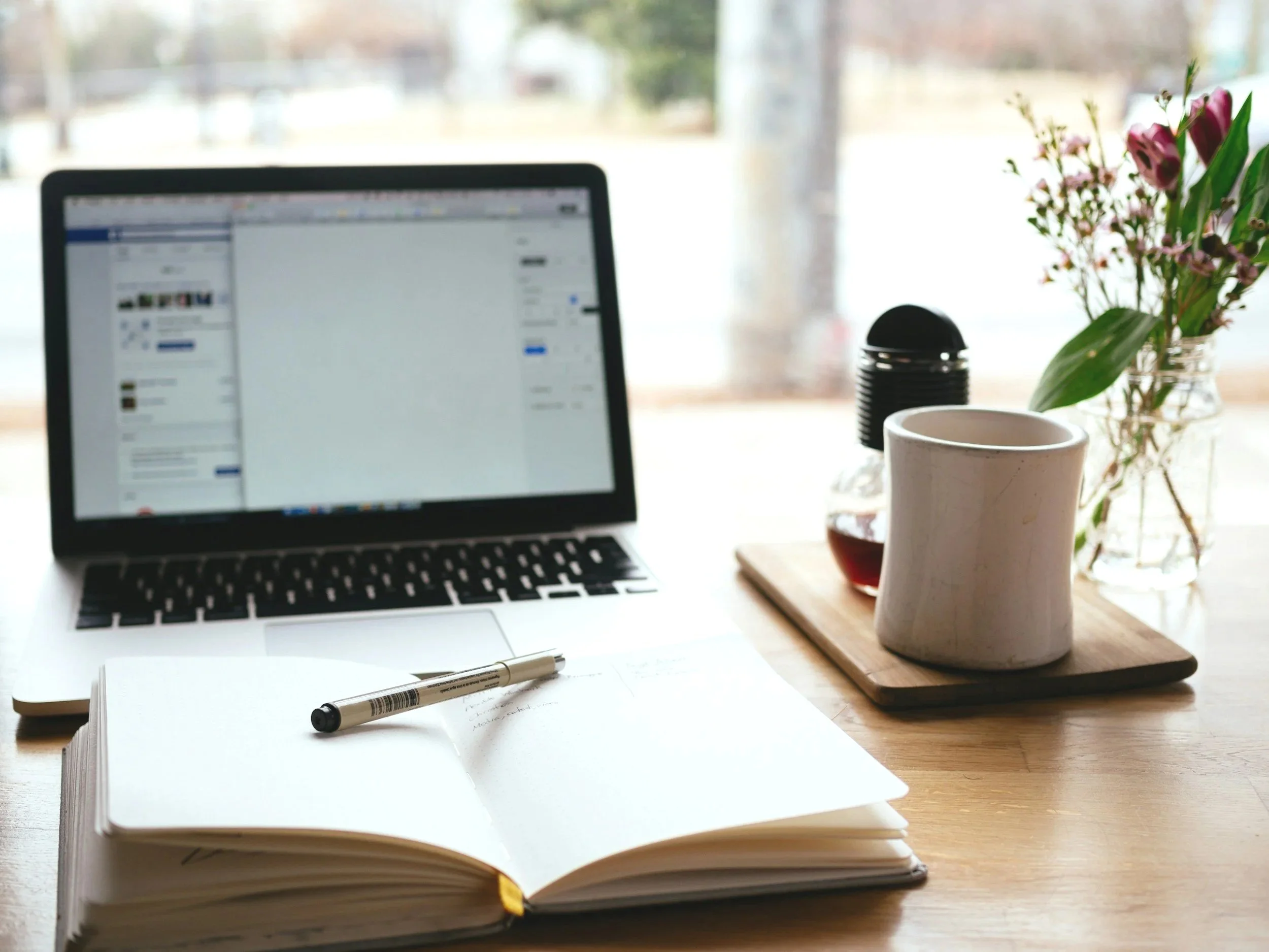A freelance technical writing and editing workspace with a laptop, an open notebook with a pen on it, a white mug, a jar of flowers, and a bottle of tea on a wooden table near a window.