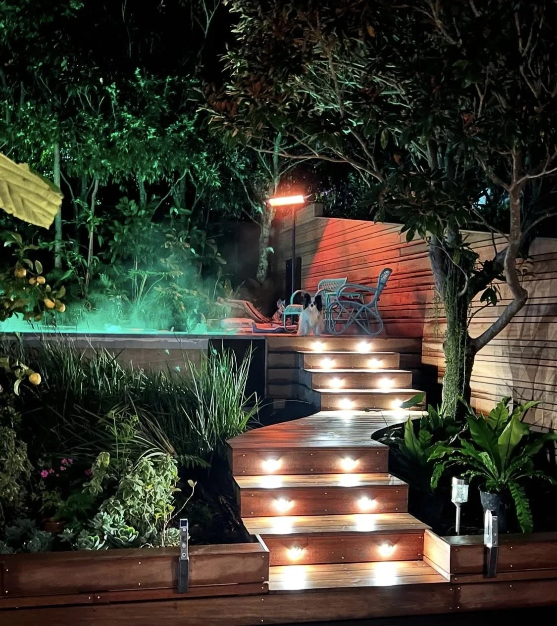 Nighttime garden scene with illuminated wooden steps leading to a patio area, a dog sitting on the steps, surrounded by lush plants and trees, with outdoor chairs, a table, and a tall lamp on the patio