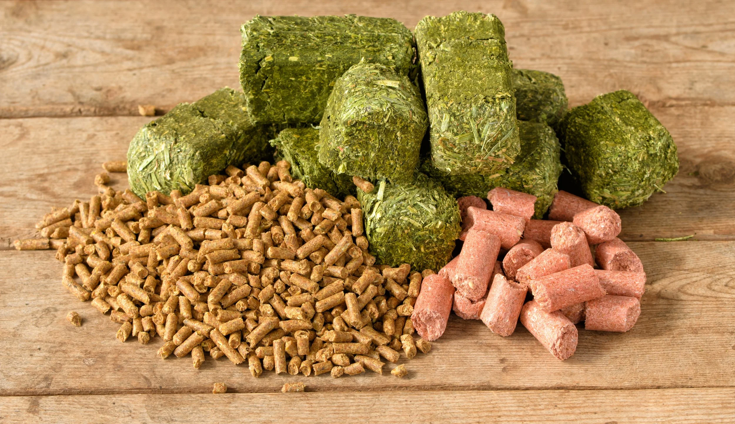 Various types of pet food including green herb-based treats, pink meaty treats, and brown pellet-based kibble, arranged on a wooden surface.