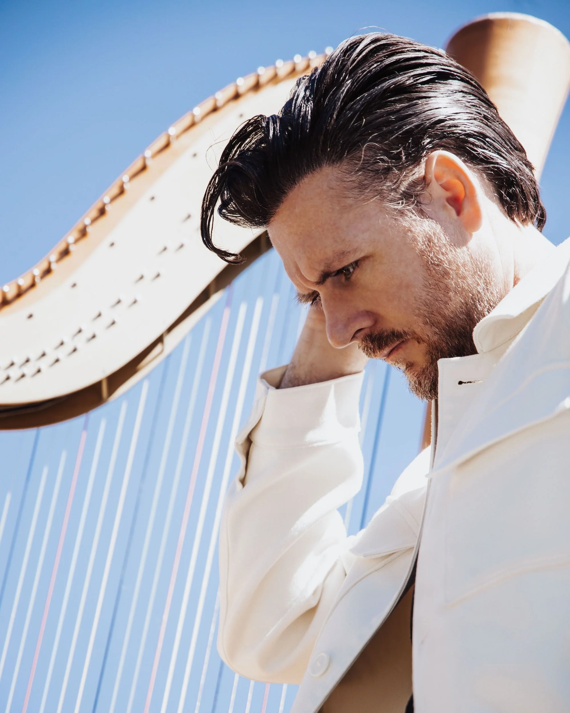 Man with dark, slicked-back hair and beard, wearing a white shirt, standing outdoors against a bright blue sky and a structure with vertical lines