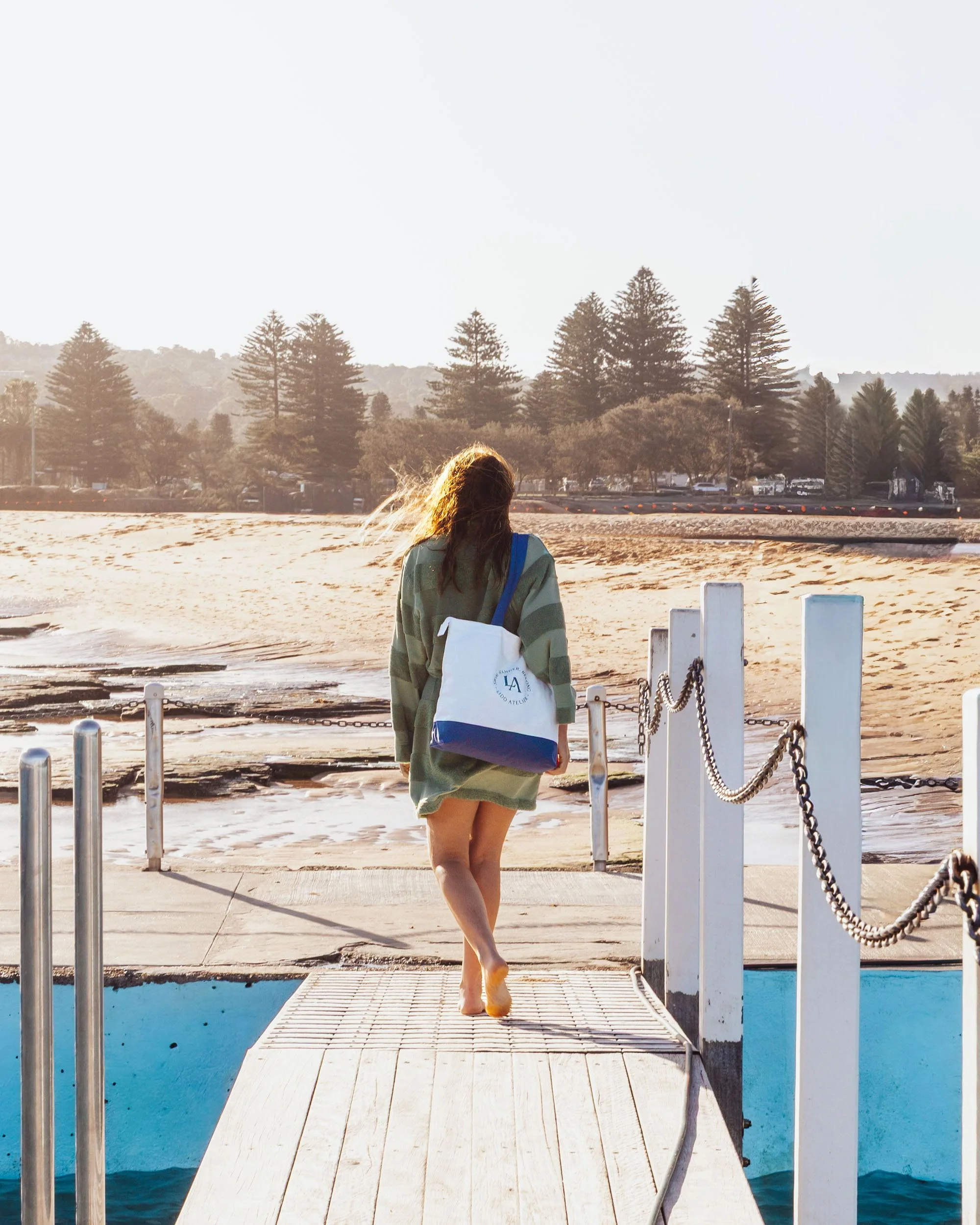 A woman with long hair, wearing a green oversized sweater and carrying a white and blue tote bag, walks barefoot off a dock towards a sandy beach and the ocean during sunset.