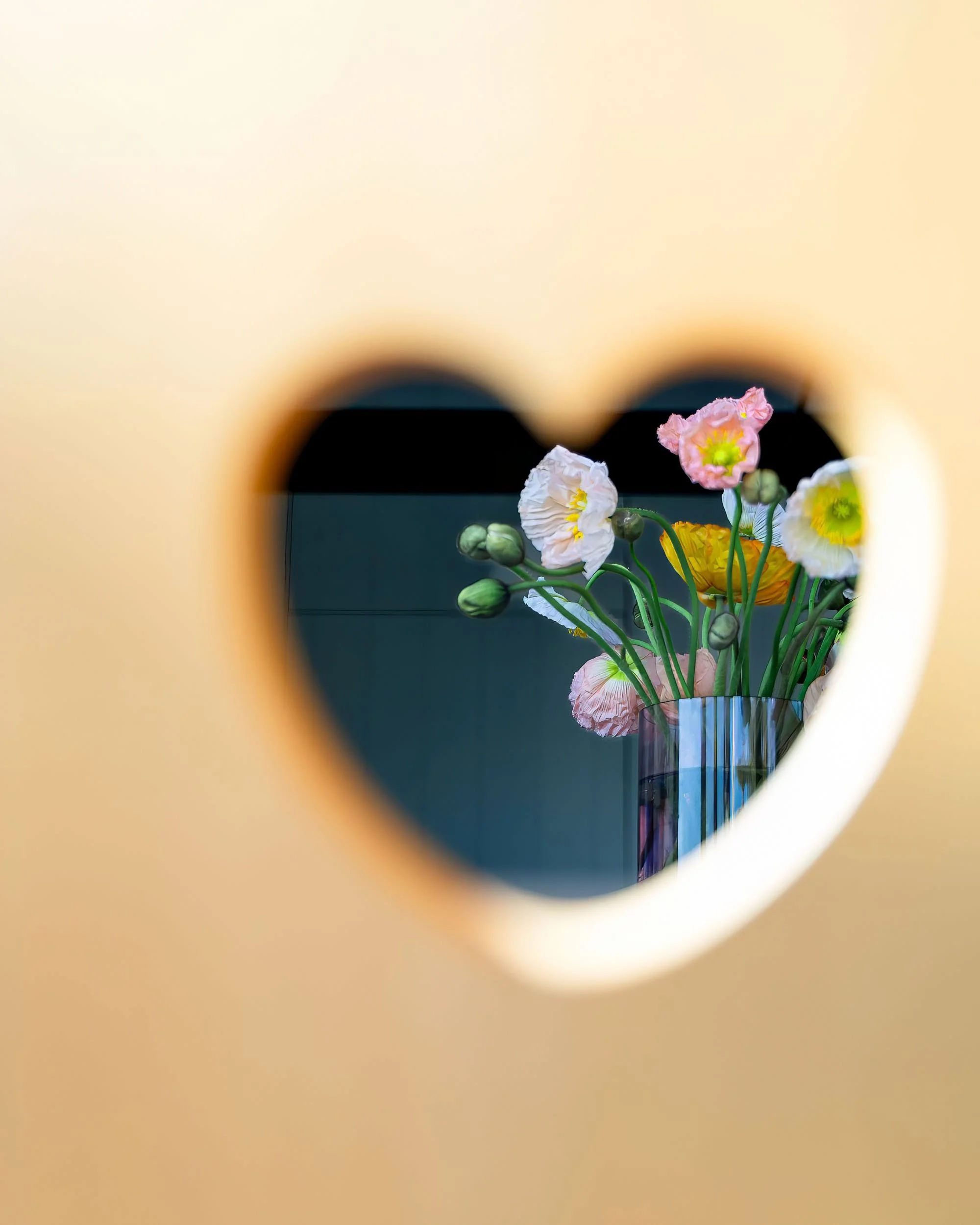 View of a bouquet of pink, white, yellow, and orange flowers seen through a heart-shaped opening in a yellow surface.