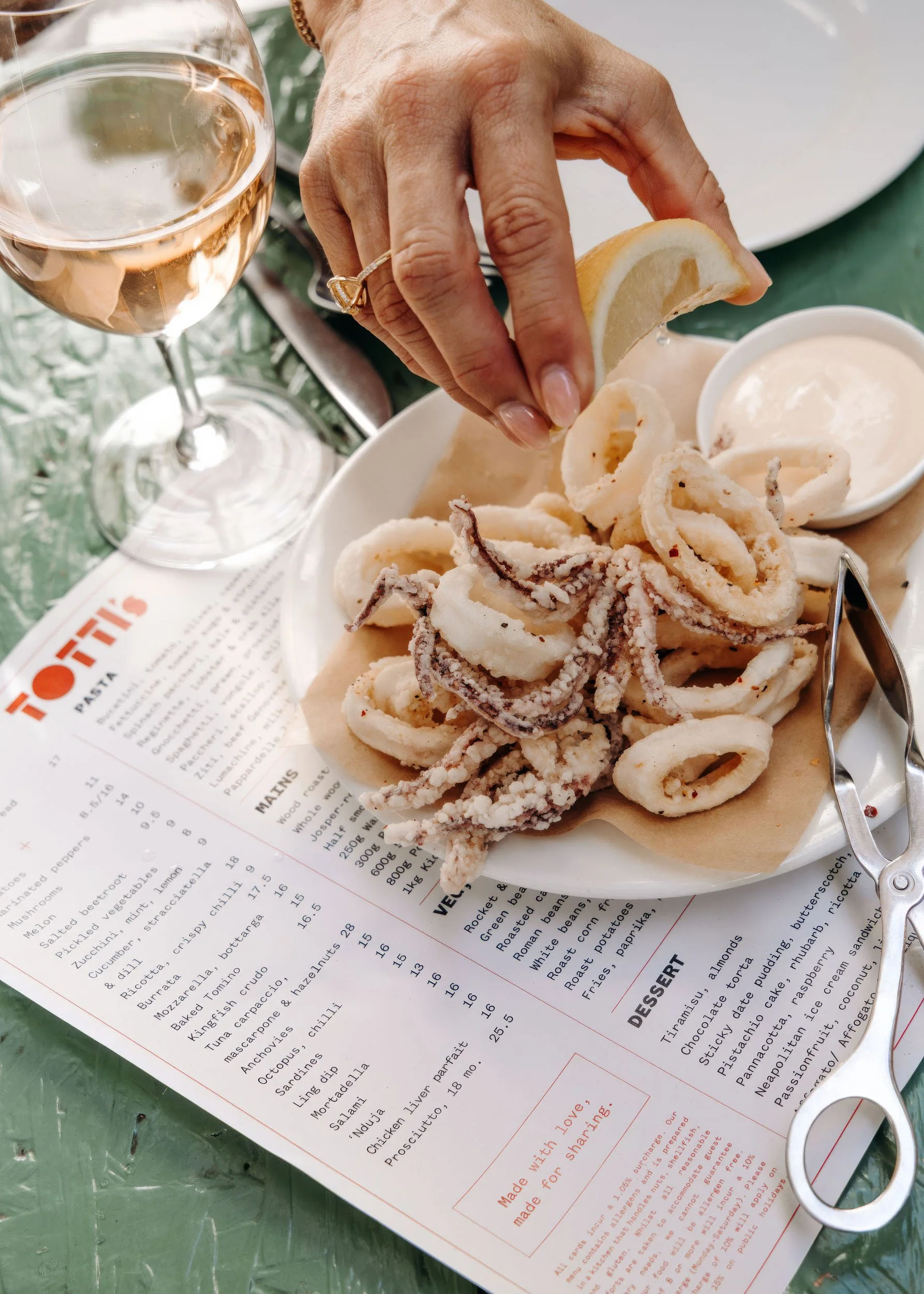 A plate of fried calamari with lemon wedges, a small dipping sauce, a glass of rosé wine, and a restaurant menu on a green table.