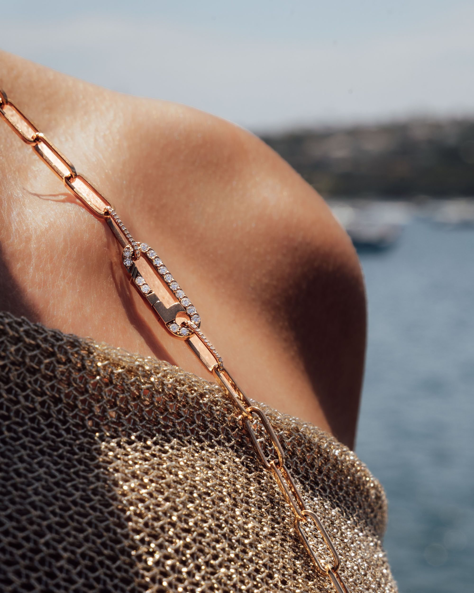 Close-up of a gold chain necklace with diamond accents draped over a person's shoulder with a blurred ocean and rocky coastline in the background.