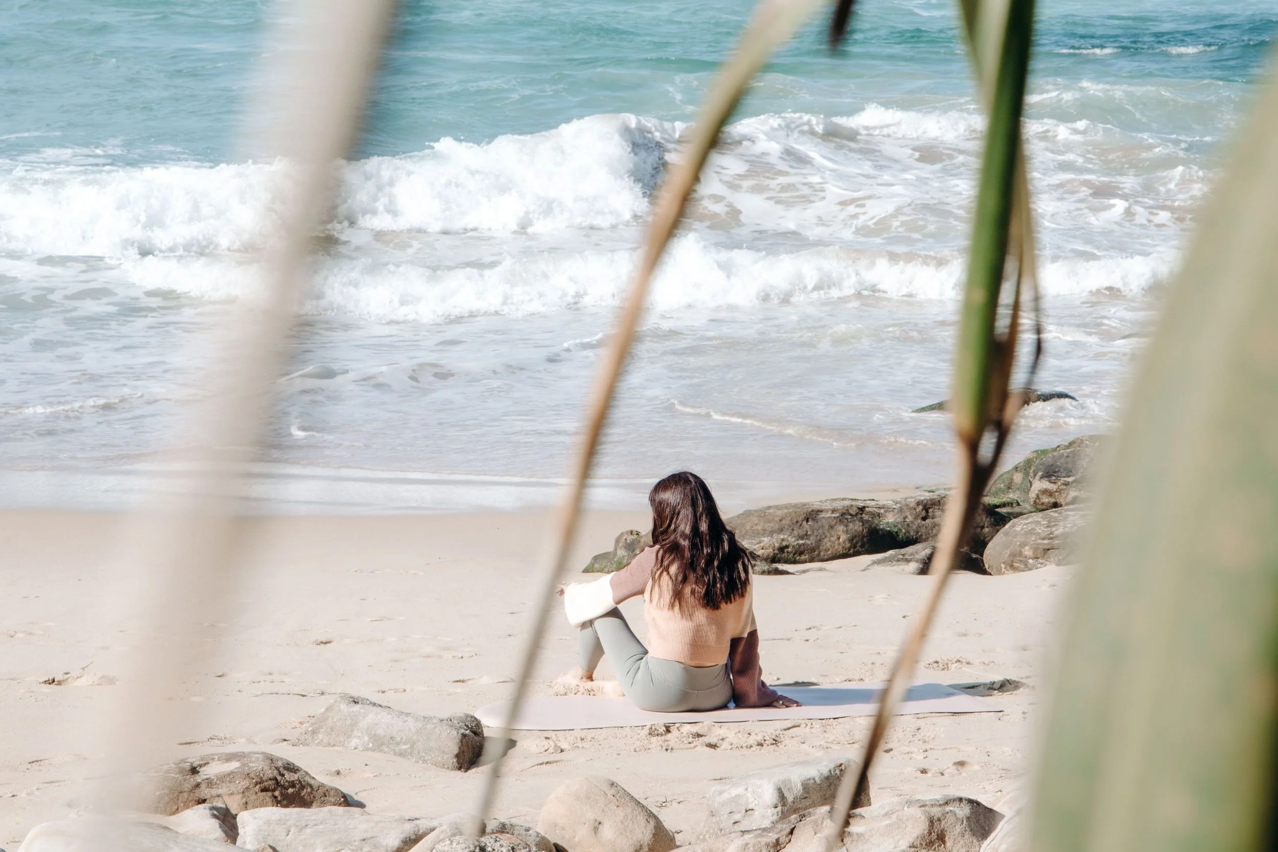 A woman with long dark hair sitting on a beach surrounded by rocks, looking at the ocean waves.