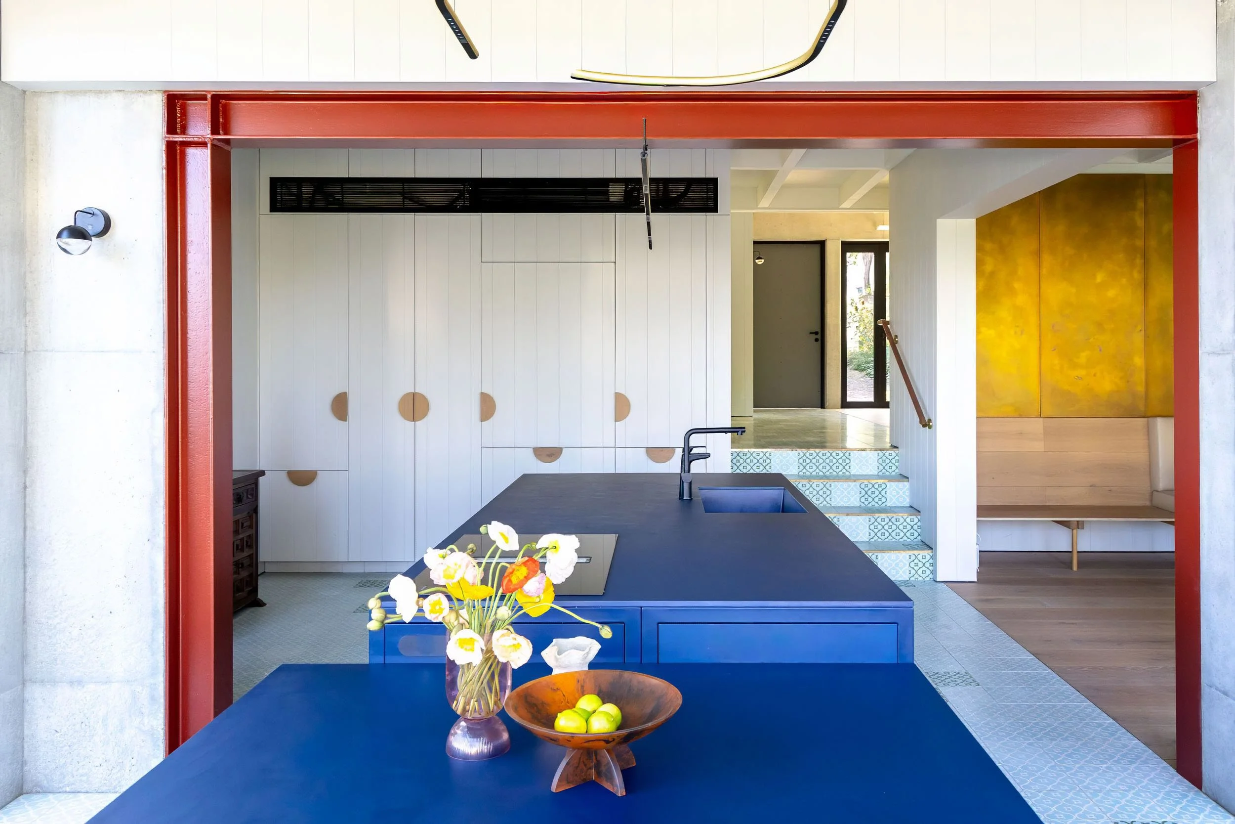 Interior view of a modern kitchen with a blue island counter, a vase of white and yellow flowers, a wooden bowl of green apples, and a staircase with patterned tiles leading to an entry door.