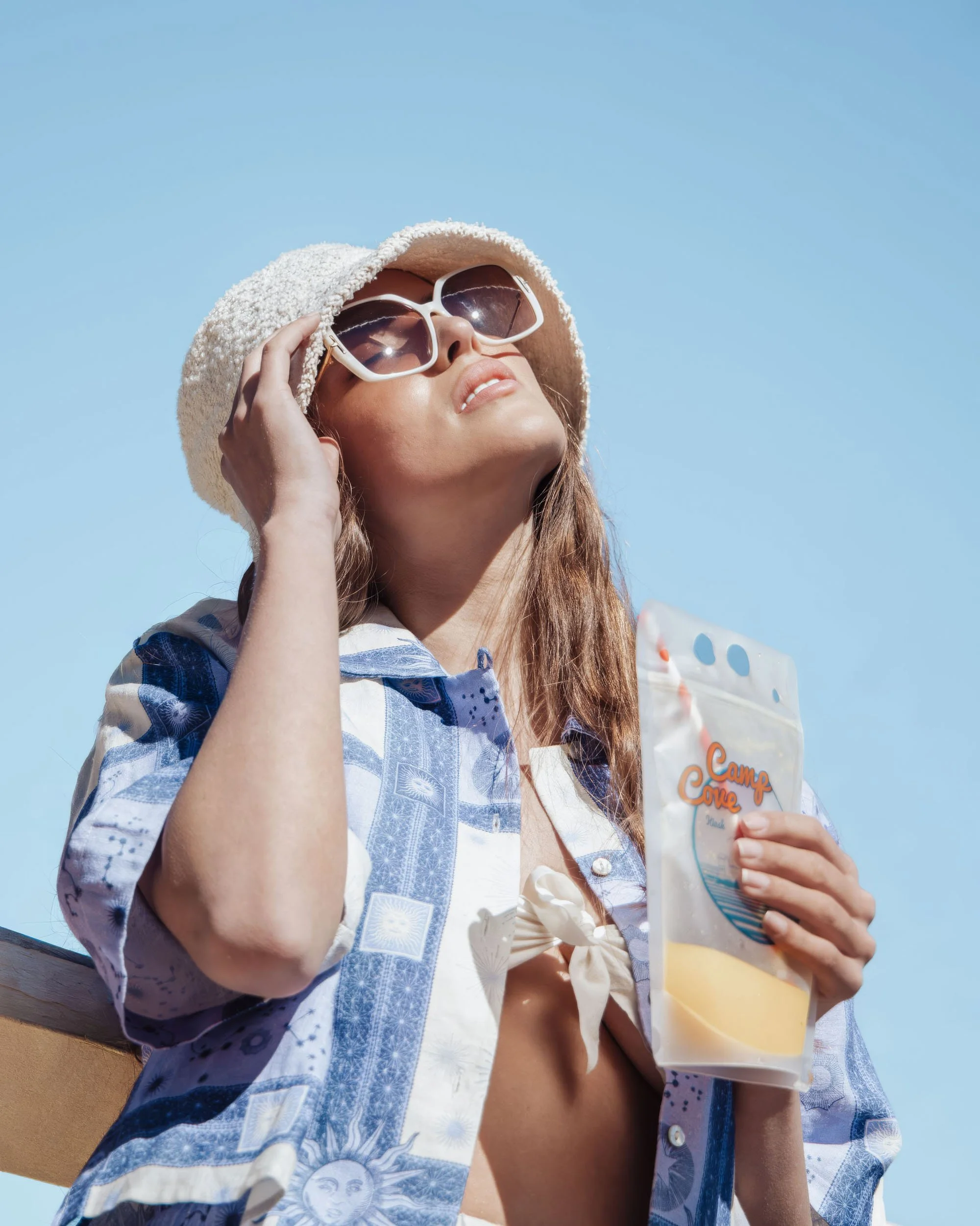 Woman wearing sunglasses and a beige sunhat, holding a bag of Camp Cove water, enjoying the sunny outdoor day.