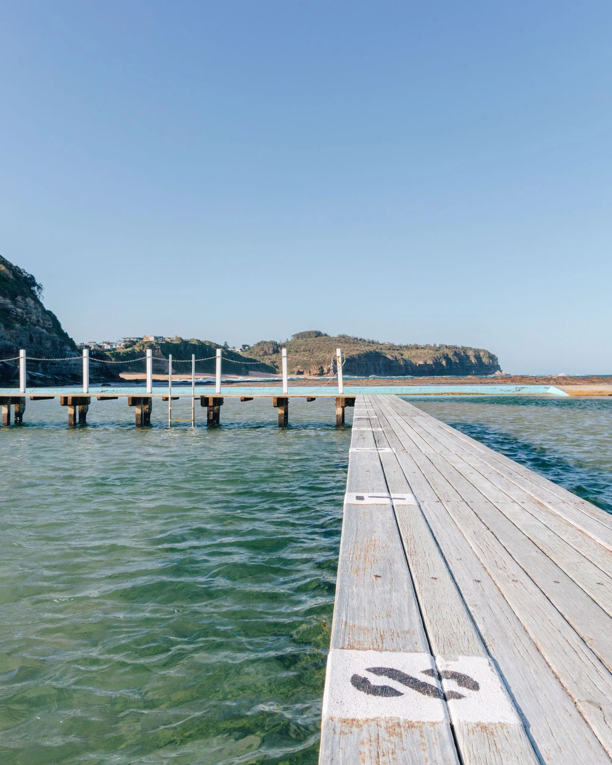 A wooden dock extending into clear, greenish water with a rocky shoreline and hills in the background under a blue sky.