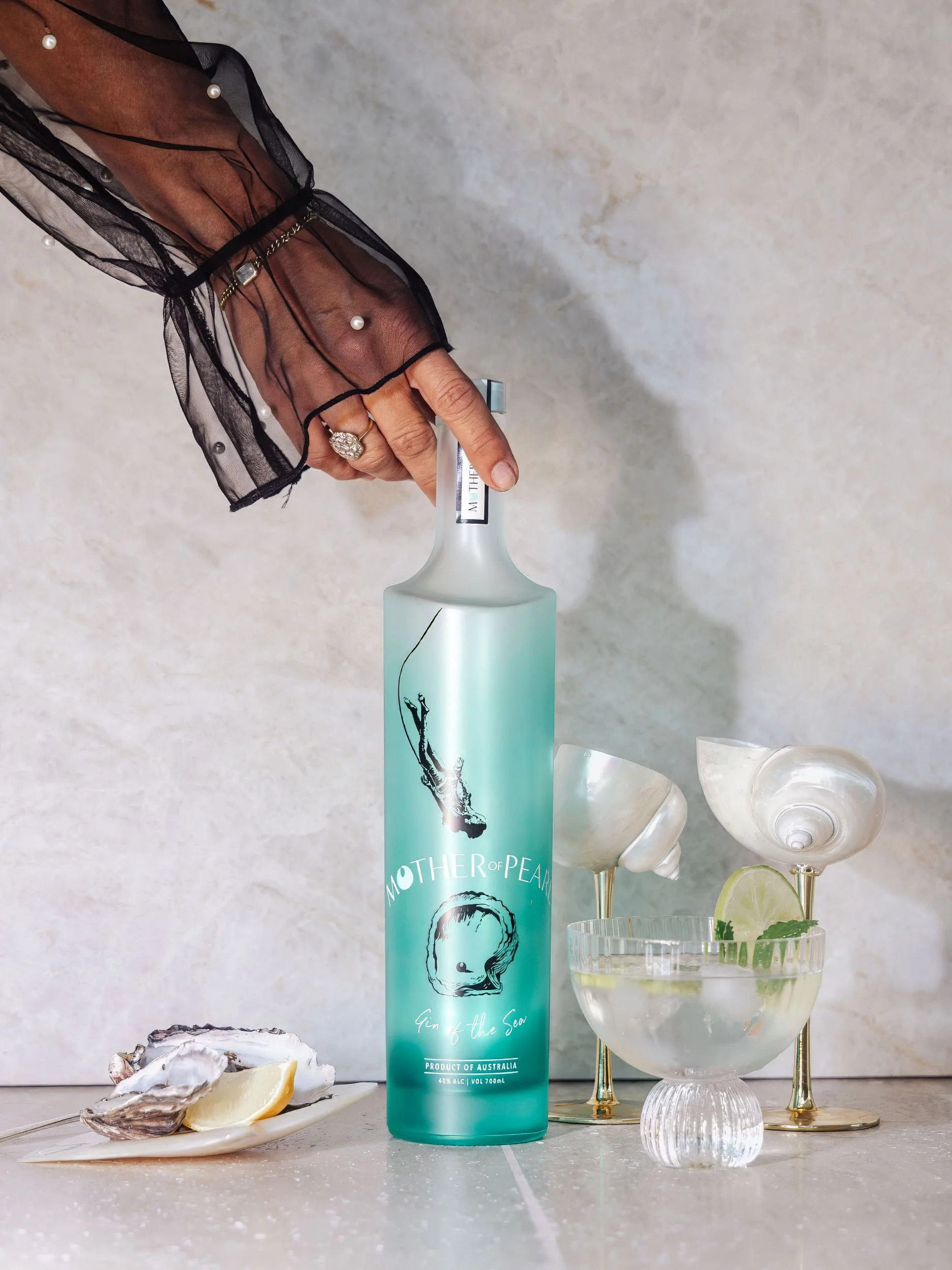 A hand with a black sheer sleeve and pearl accents holding a bottle of Mother of Pearl Gin of the Sea, surrounded by oyster shells with lemon, and cocktail glasses with lime and mint, on a white surface.