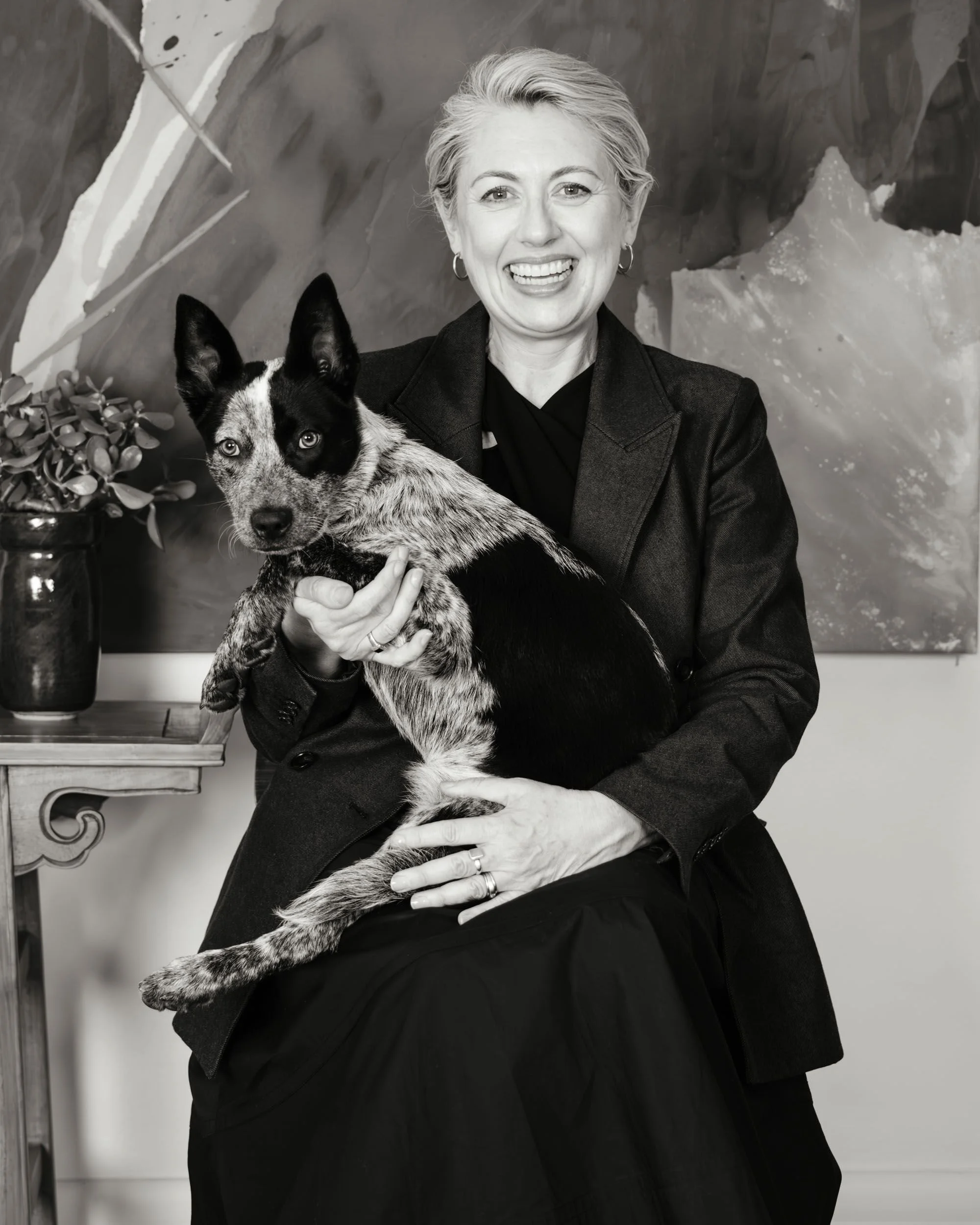 A woman with short, light-colored hair holding a black and white dog with blue eyes inside a room with a decorative painting and a potted plant.