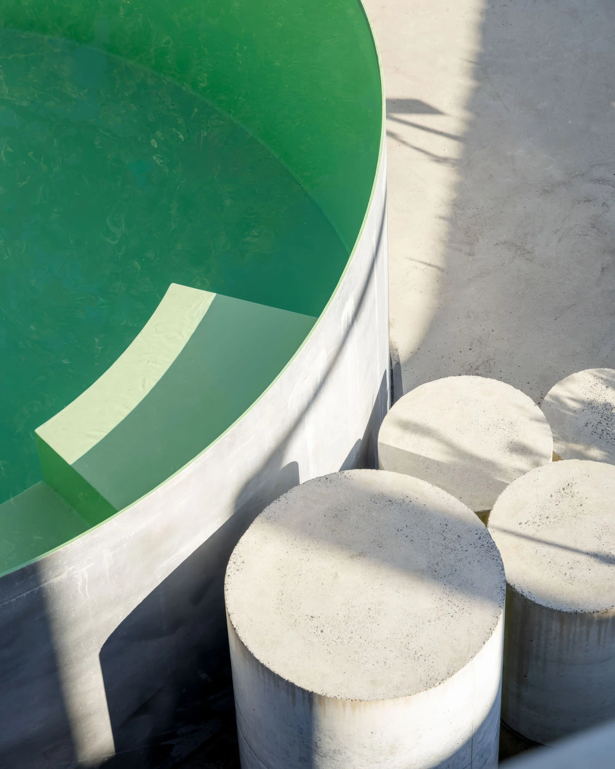 Close-up of a modern outdoor sculpture with a large green circular surface and three white cylindrical concrete bases, casting shadows on a concrete ground.