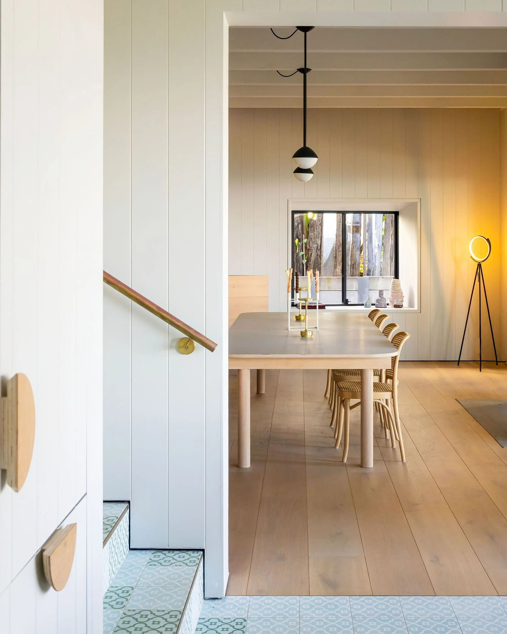 View of a modern dining room with a light wooden table, beige chairs, and a window with outdoor trees. The space features a white vertical paneled wall, a black pendant light fixture, and a yellow floor lamp with a circular light, illuminated in the 