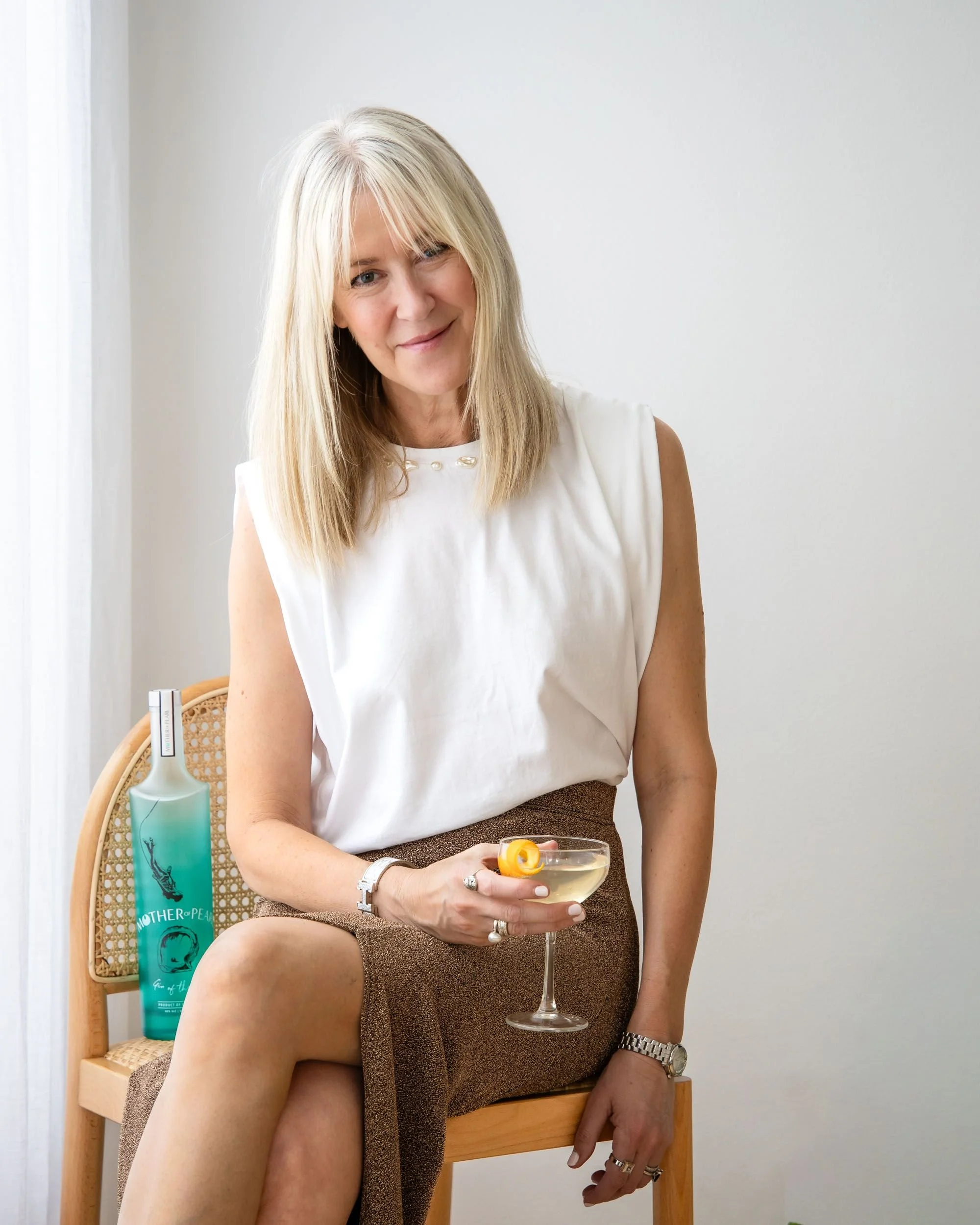 A woman with blonde hair wearing a white sleeveless blouse and brown skirt sitting on a wooden chair, holding a martini glass with a lemon twist, with a bottle of liquid on the chair beside her.