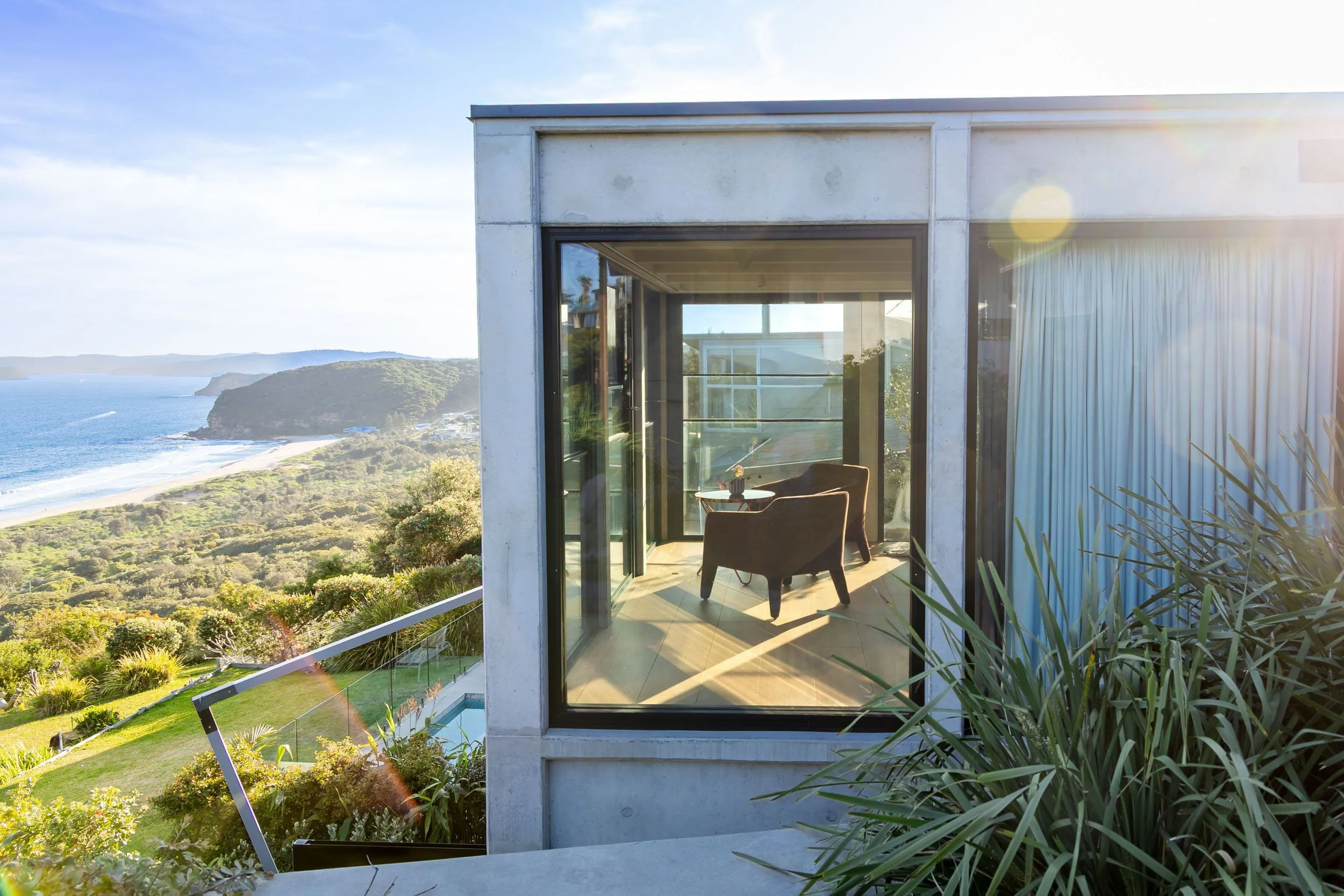 Cozy balcony with two chairs and a small table, overlooking a scenic coastal landscape with ocean, hills, and greenery, illuminated by bright sunlight.