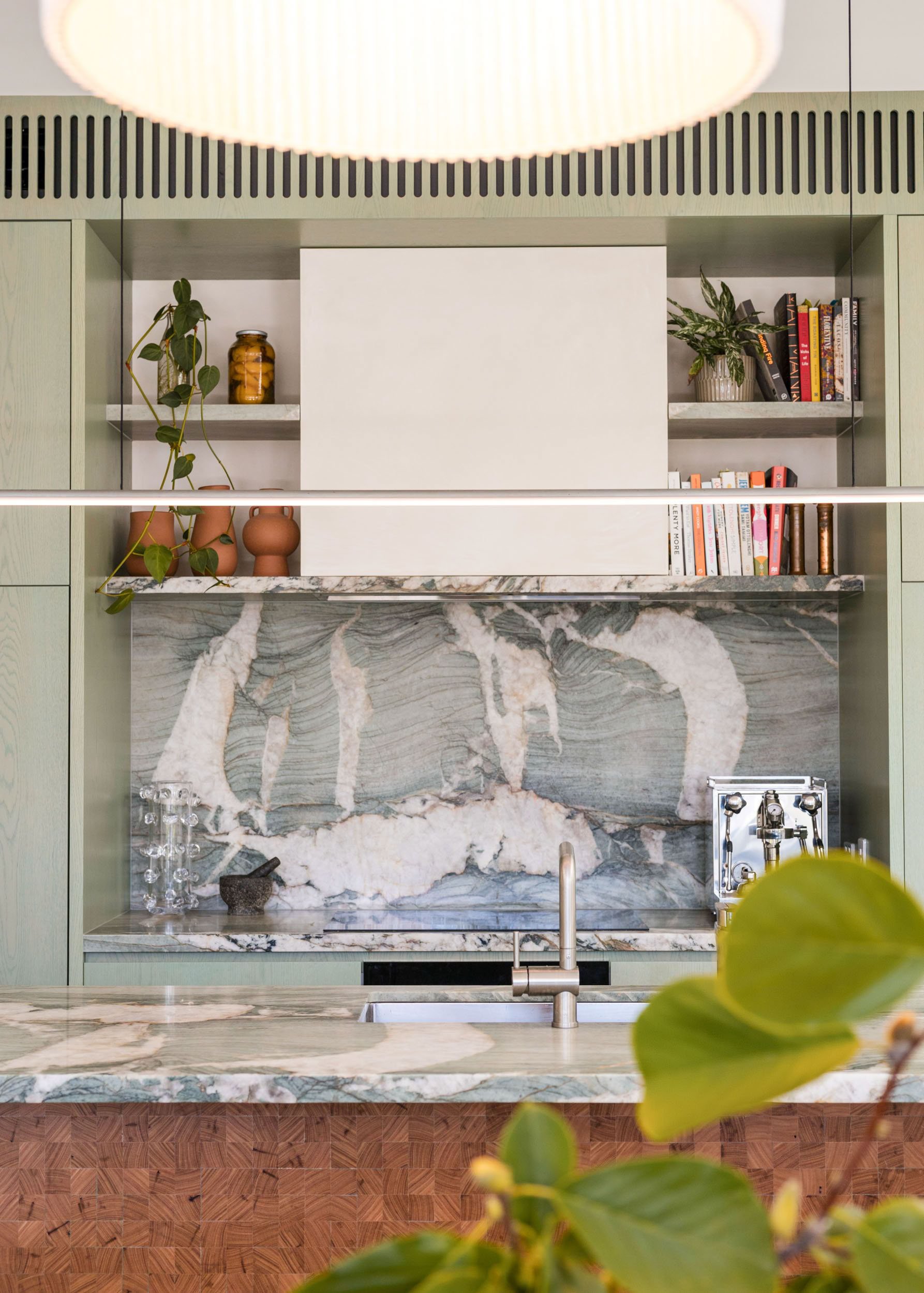 A modern kitchen with green cabinets, a marble backsplash, and a marble countertop. There are open shelves with books and plants, and a decorative jar. The foreground shows some green leaves.
