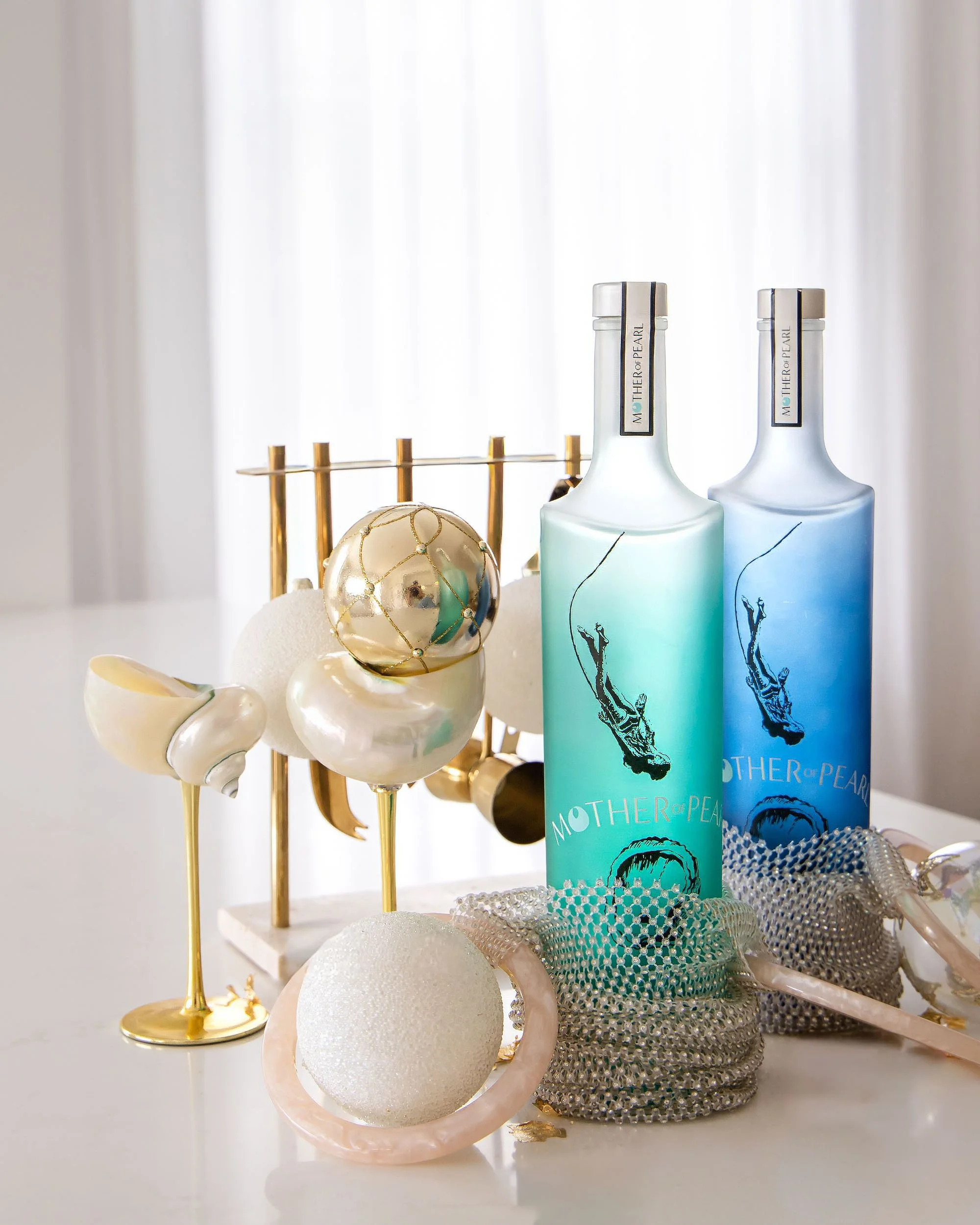 Decorative items including a gold and white wire globe, white and pearl-colored spheres, a gold and white stand, two bottles of Mother of Pearl liquor with ocean-themed labels, and a pink shell-shaped container with a white textured ball inside, arra