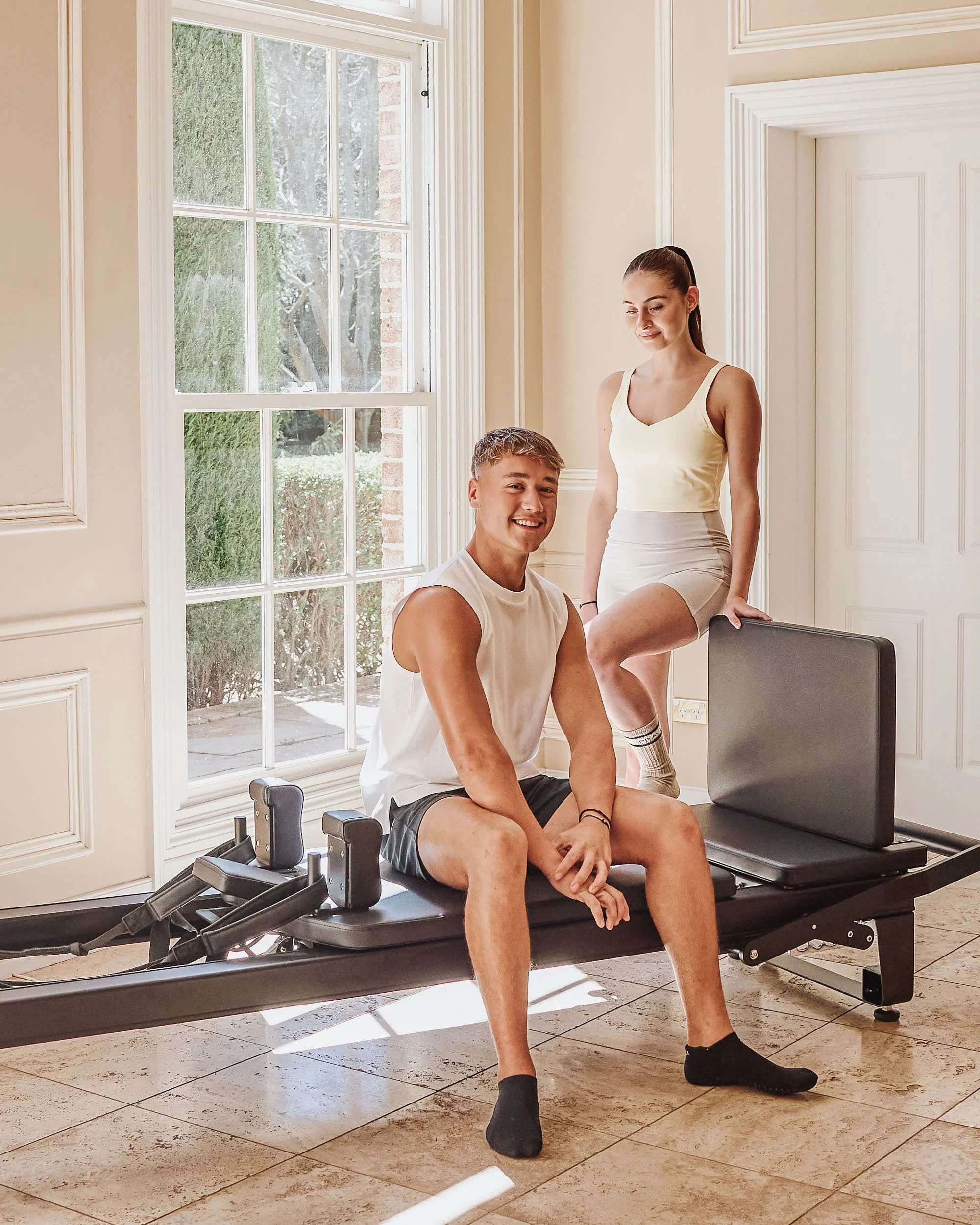 A young man and woman in athletic workout clothes indoors with sunlight coming through large windows. The man is sitting on a Pilates reformer machine, smiling at the camera, while the woman is standing next to him, leaning on the machine with one fo