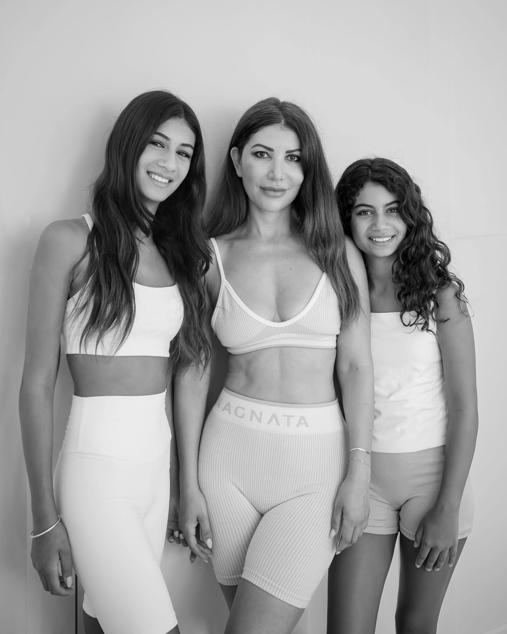 Three women standing together, wearing sports bras and shorts or leggings, smiling, against a plain background.