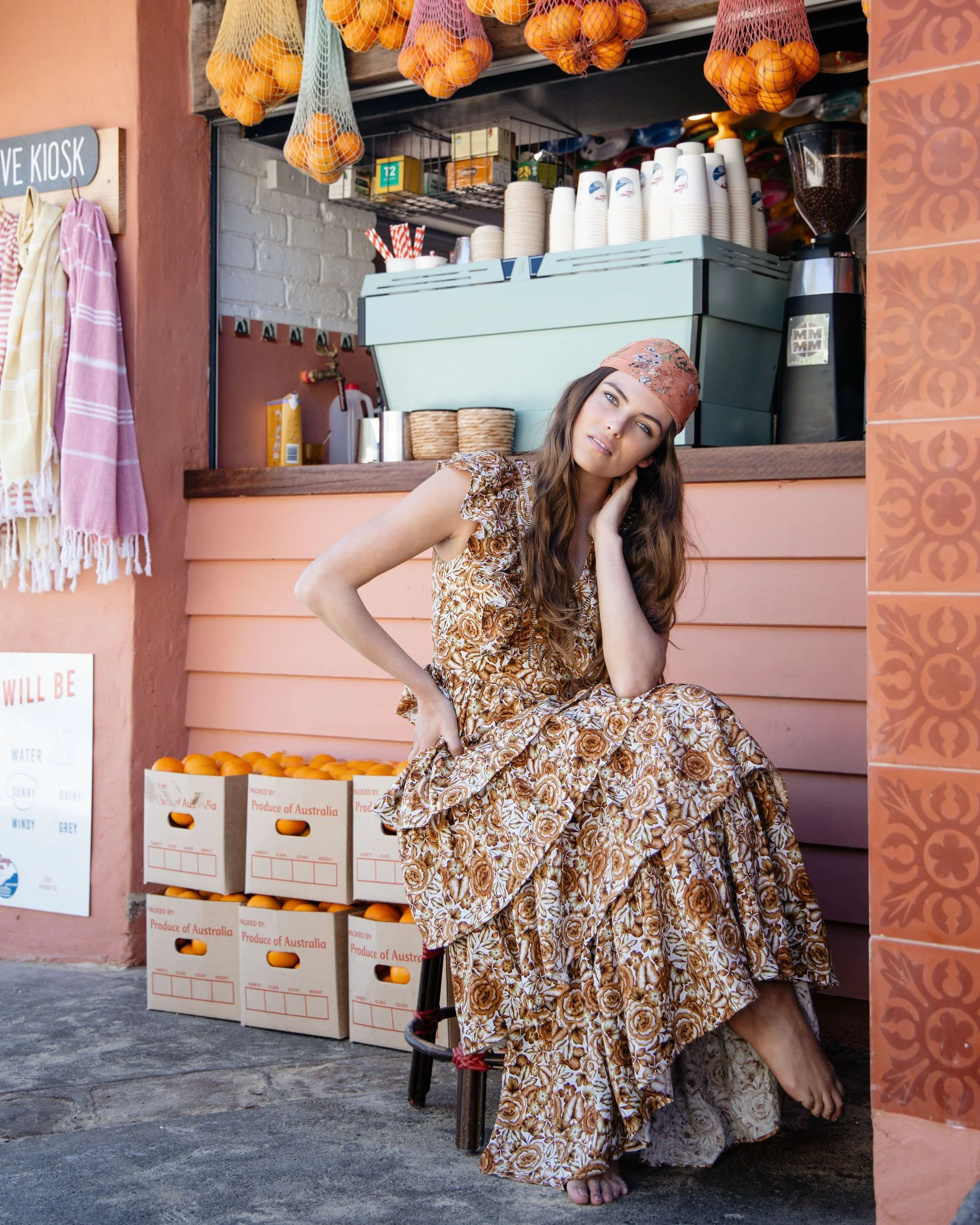 A woman with long brown hair wearing a floral dress and headscarf sitting outside a fruit stand, with oranges in crates and various items in the background.