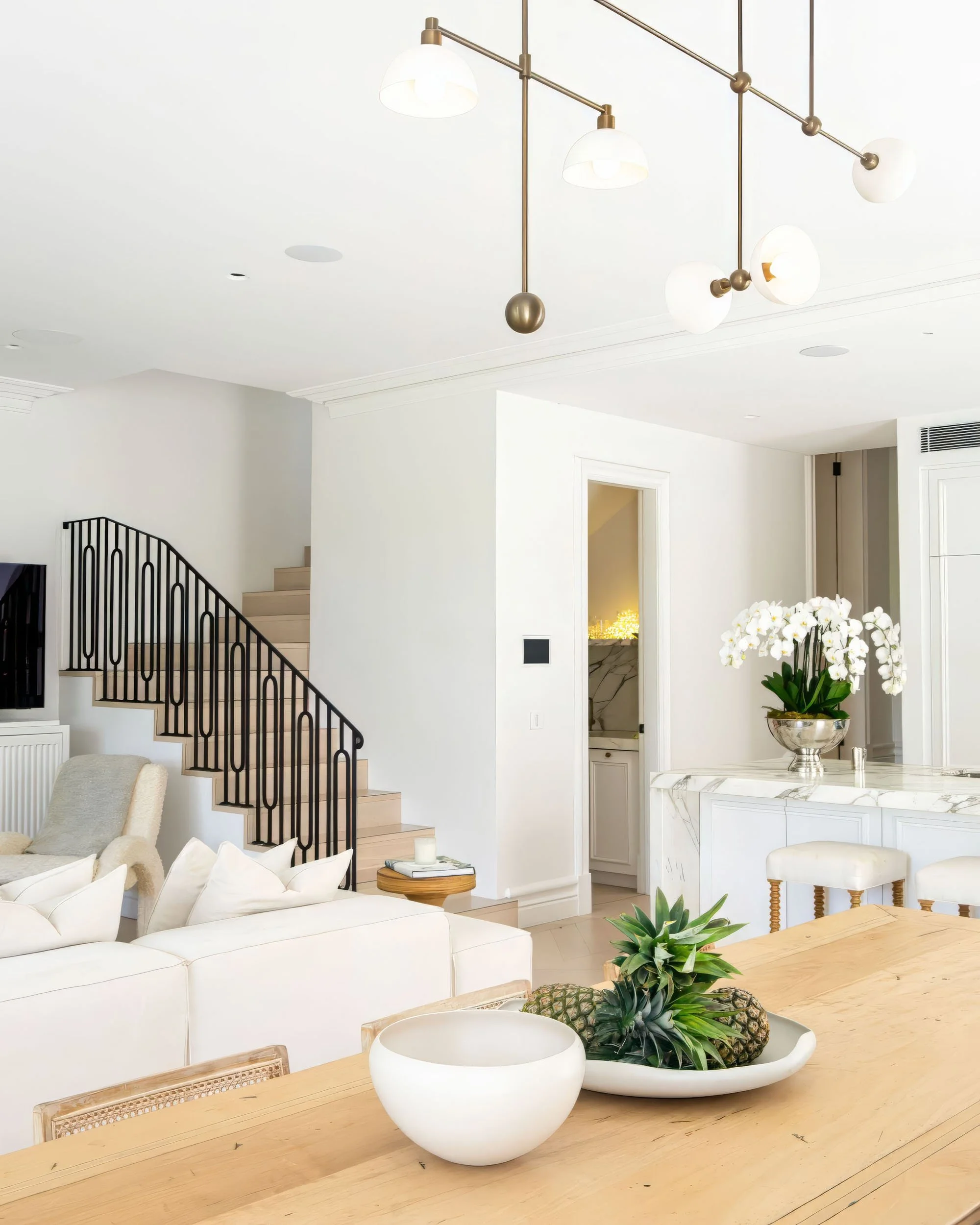 Modern living room with white walls, a staircase with black railing, a wooden dining table with a bowl of pineapples, a white sofa, and a contemporary chandelier with globe lights.