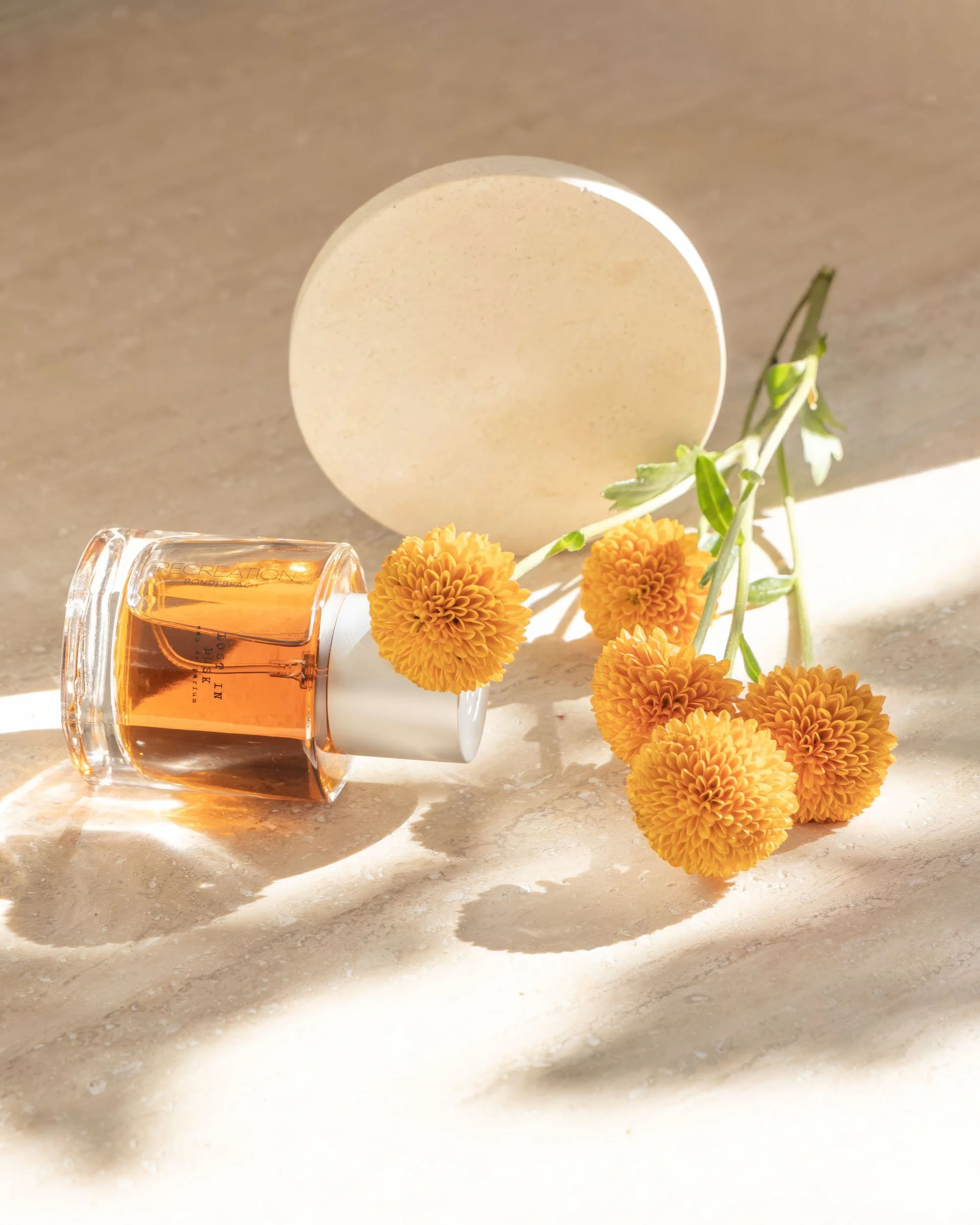 A bottle of amber-colored perfume lying on a light surface next to a small bunch of yellow-orange flowers and a white round container.