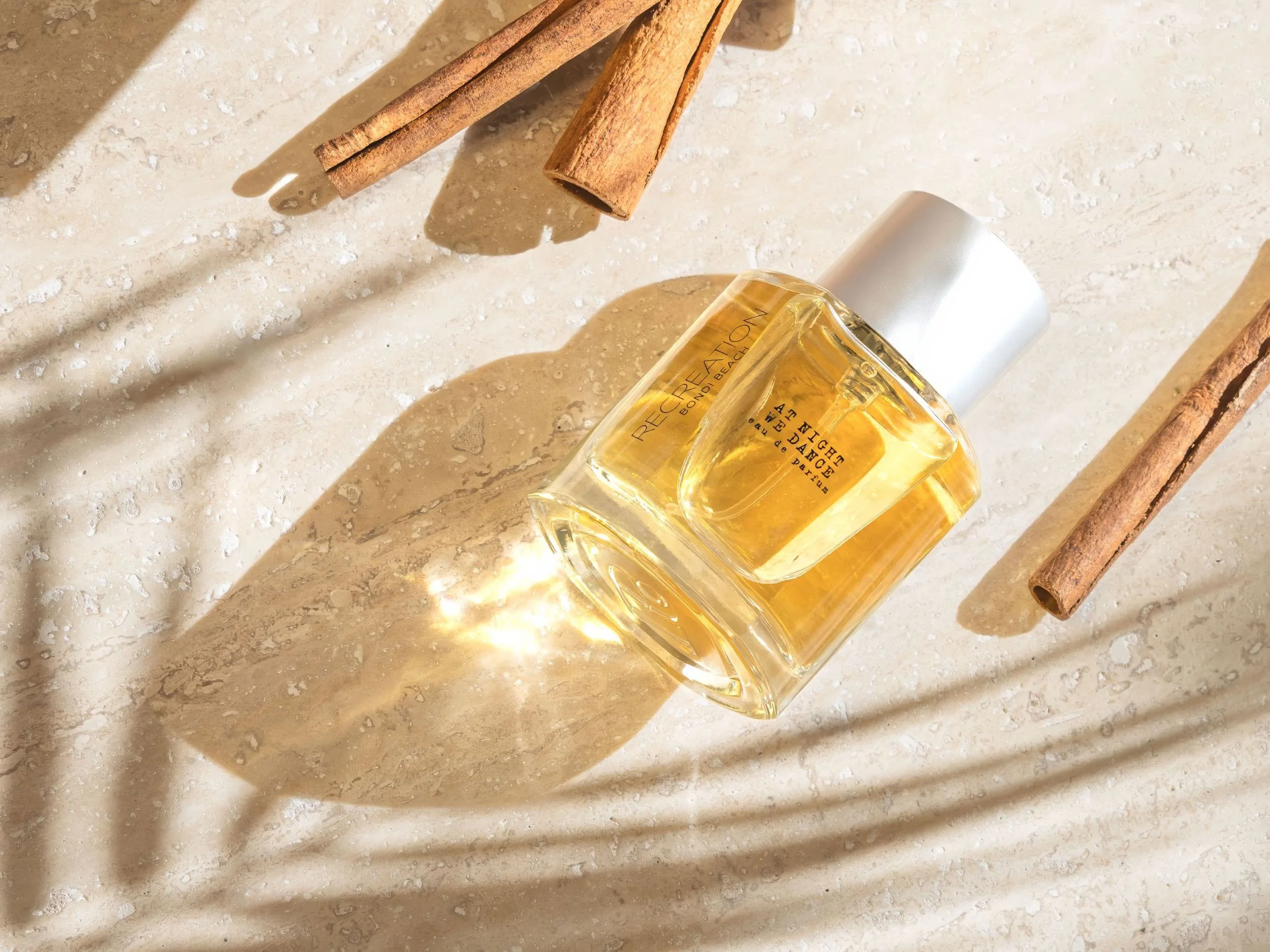A bottle of perfume labeled 'Recreation Bodhi Beach' lying on a beige textured surface, with cinnamon sticks casting shadows nearby.