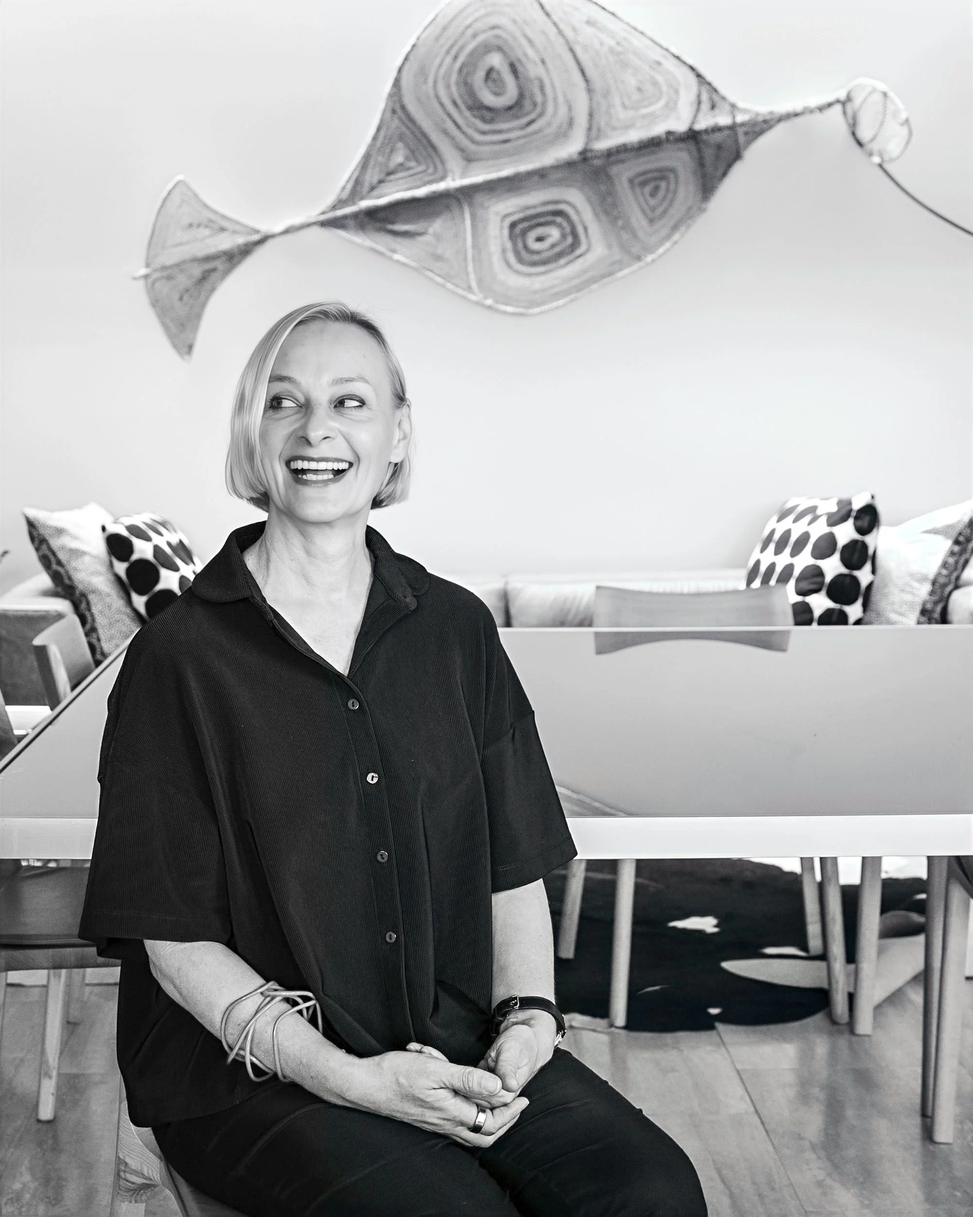 A woman with short blonde hair smiling and sitting indoors, with modern art featuring a stylized fish above her, and a dining area with polka dot cushions in the background.