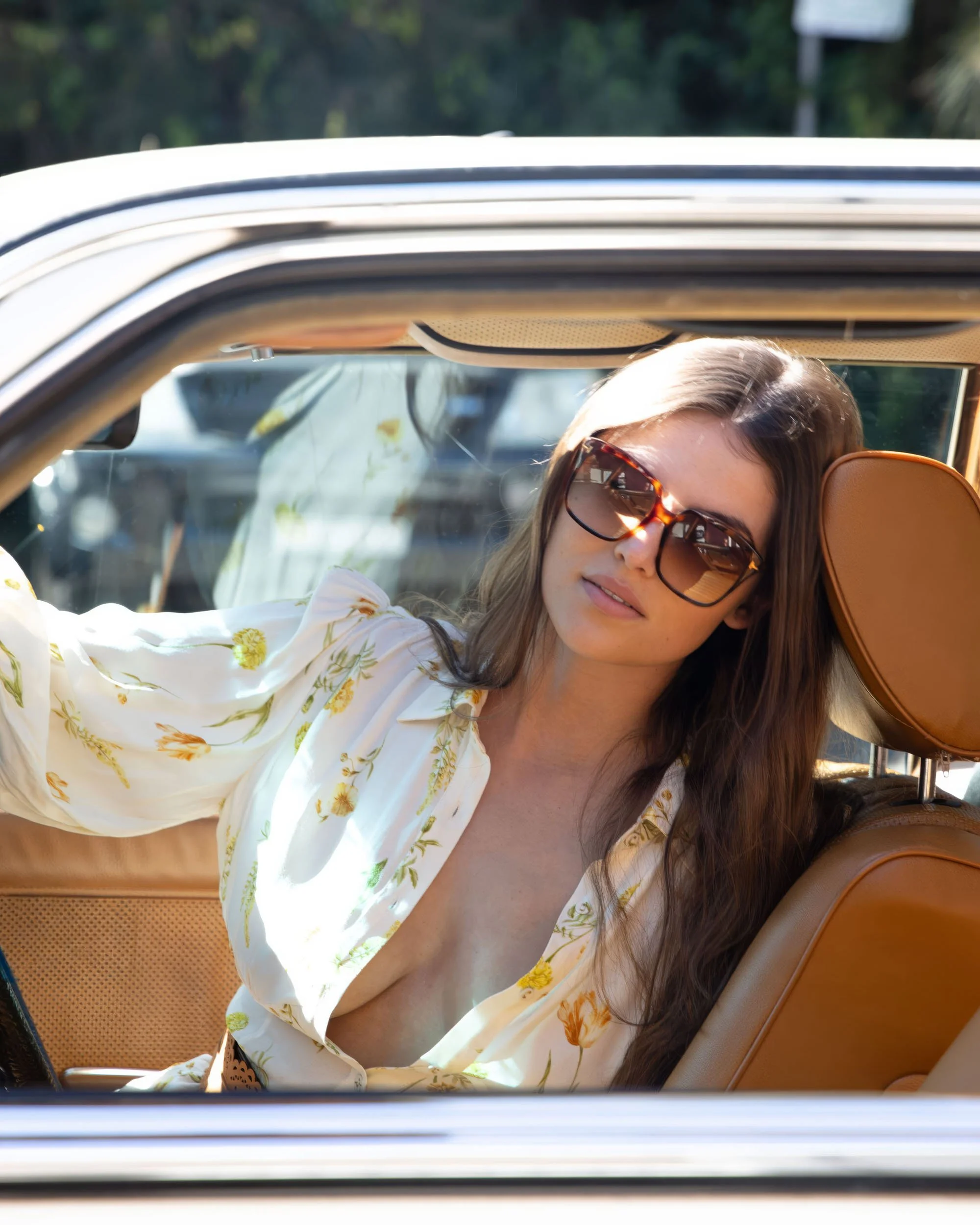 A woman with long brown hair wearing sunglasses and a floral shirt or dress, sitting in the backseat of a car with tan interior, looking out of the window.