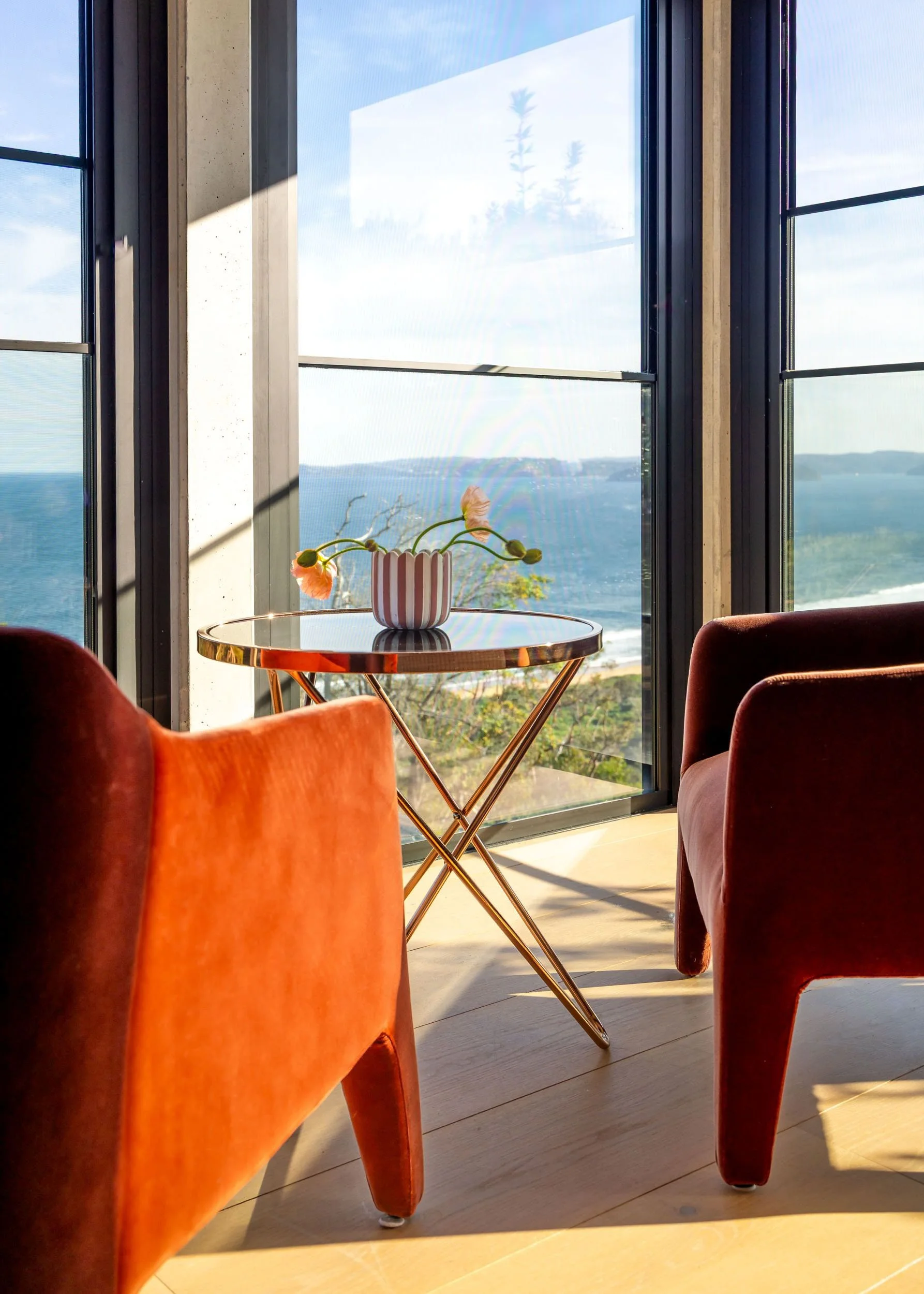 A cozy interior with two orange velvet chairs and a small round glass-top table with a striped vase and flowers inside, overlooking a large window with a view of ocean and landscape.