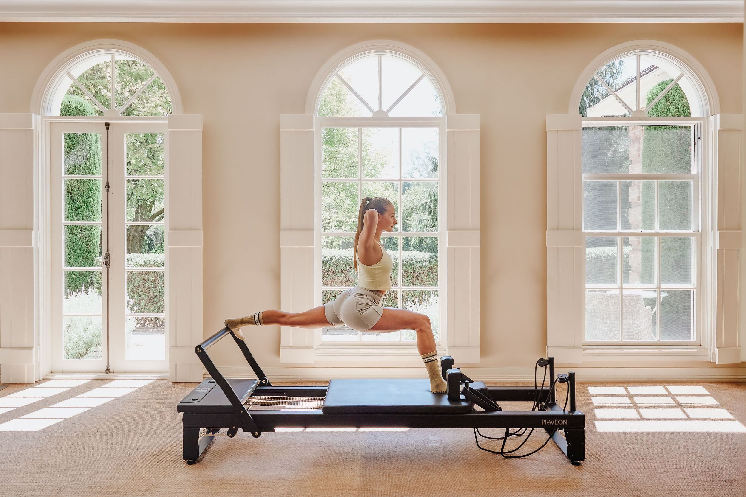 A woman in workout clothes practicing Pilates on a reformer machine in a sunny room with large arched windows.