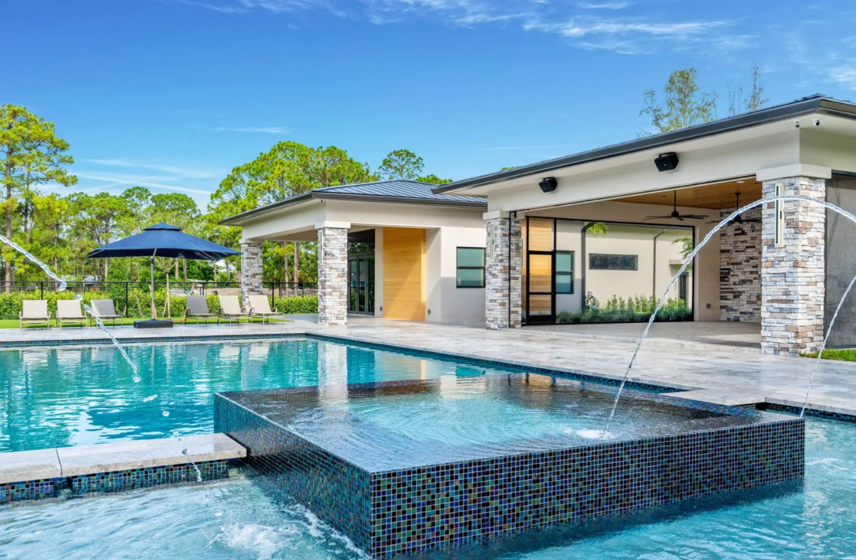 Aerial view of a modern backyard with a swimming pool, hot tub, and shaded patio areas inside a grassy yard with trees.