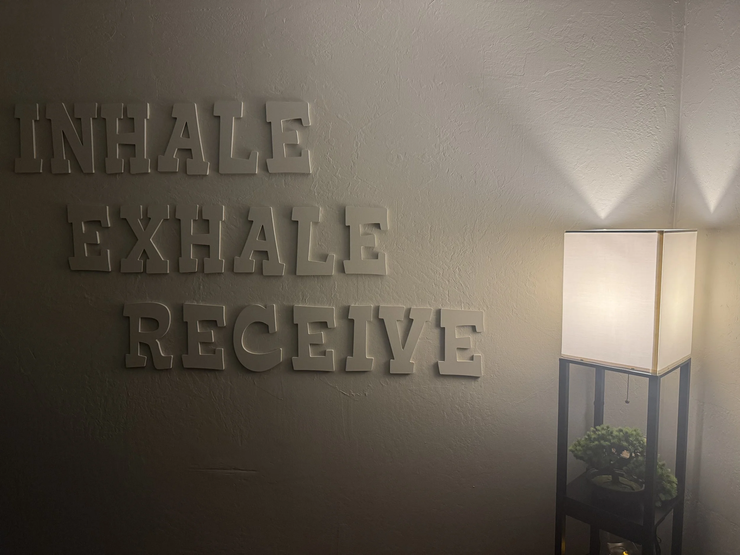 Wall decor with the words 'INHALE EXHALE RECEIVE' and a lit floor lamp with a potted plant underneath.