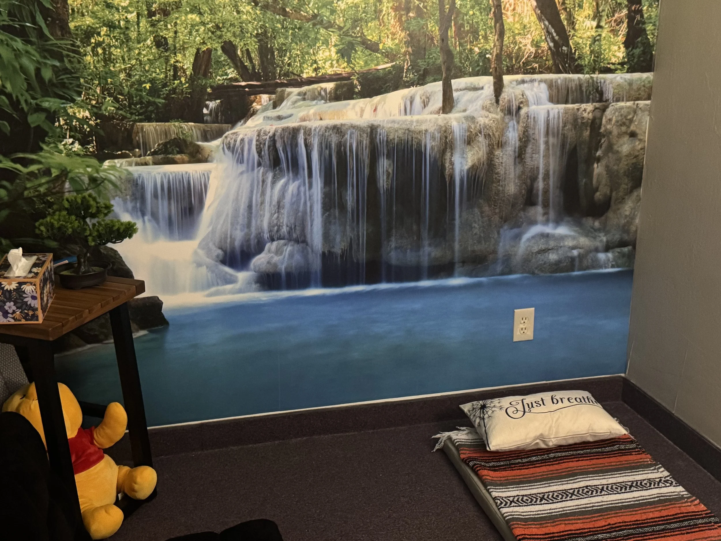 A room with a wall mural of a waterfall and forest scene. There is a small table with a potted plant, a tissue box, and a stuffed toy. On the floor, there is a pillow with the text 'Just breathe' on a folded blanket or mat.