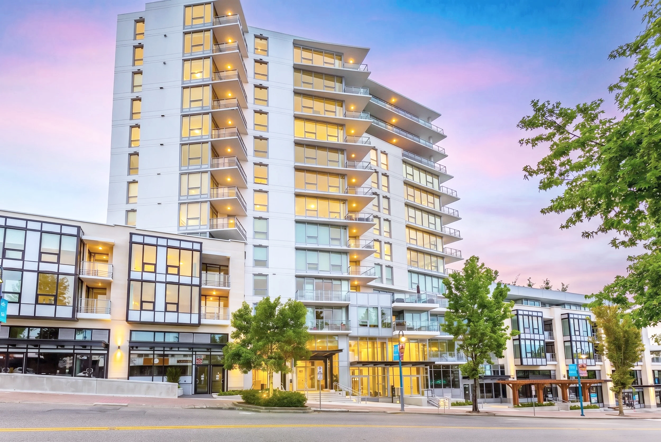 Modern multi-story residential building with large glass windows and balconies, situated in an urban area with trees and a street in front, during twilight.