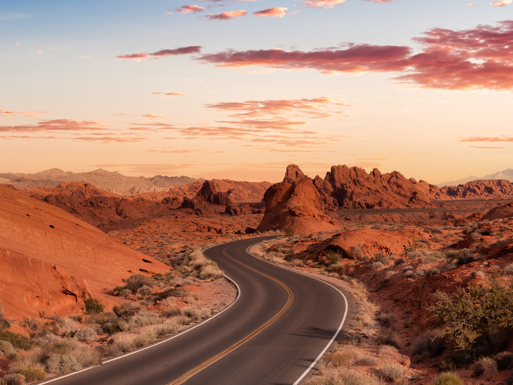 A winding road through a desert landscape with red rock formations under a pink and orange sunset sky.