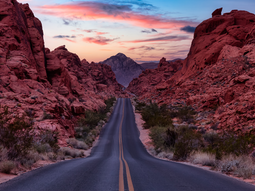 A long, empty desert road winding through red rock canyon under a colorful sunset sky.