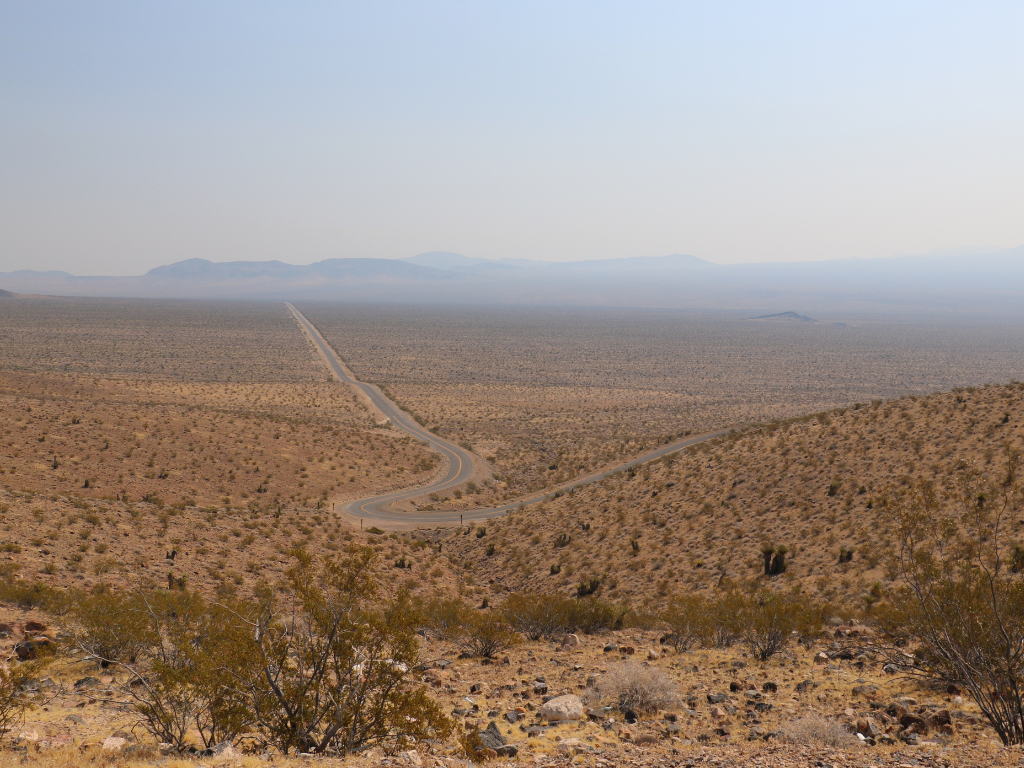 A winding road cuts through a vast, arid desert landscape with sparse bushes and distant mountains under a clear sky.