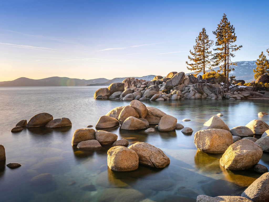 Sunset over a lakeside with large rocks and pine trees on the shore, mountains in the background