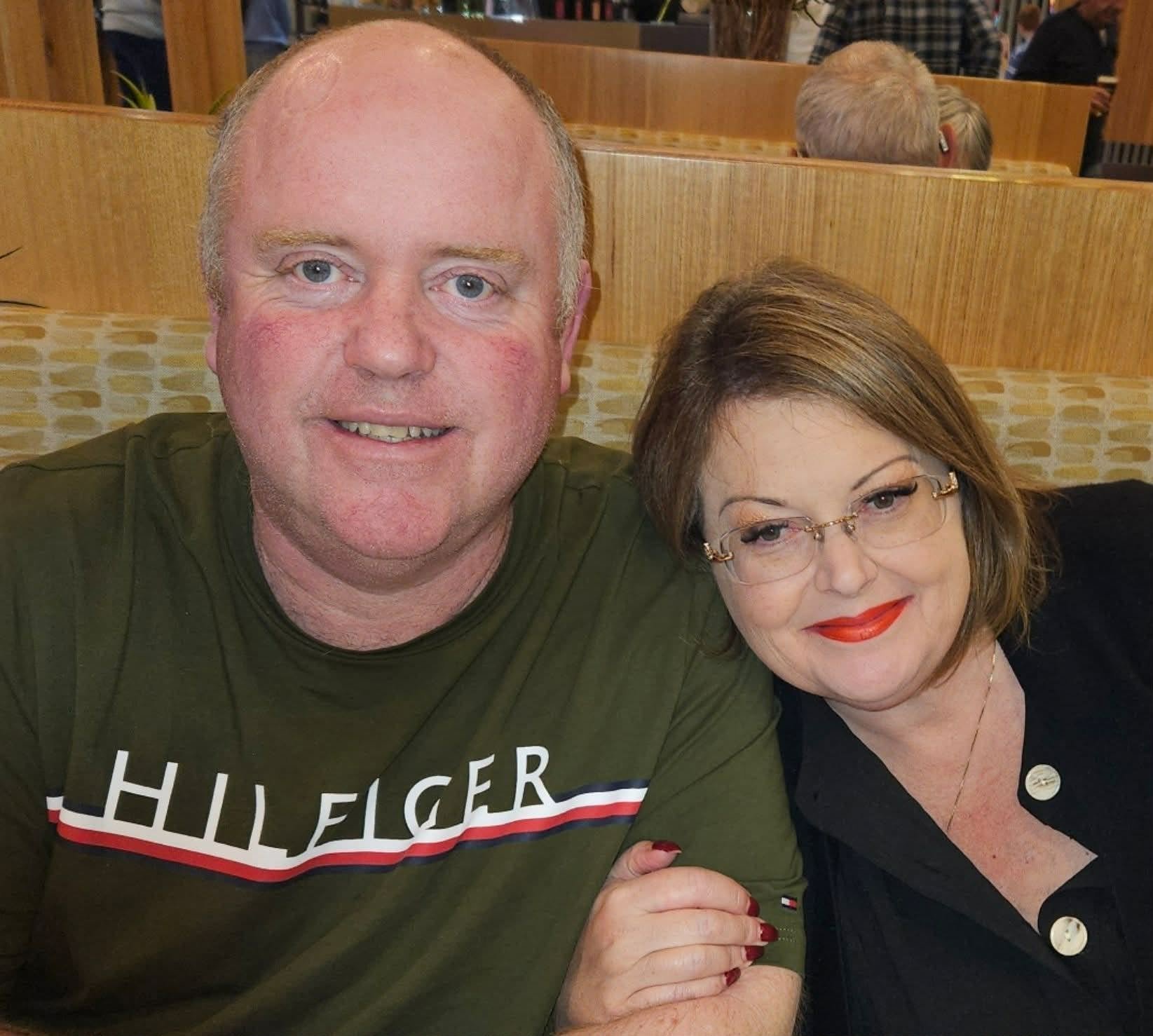 A man and woman sitting together in a restaurant, smiling at the camera, with a wooden booth and other patrons visible in the background.