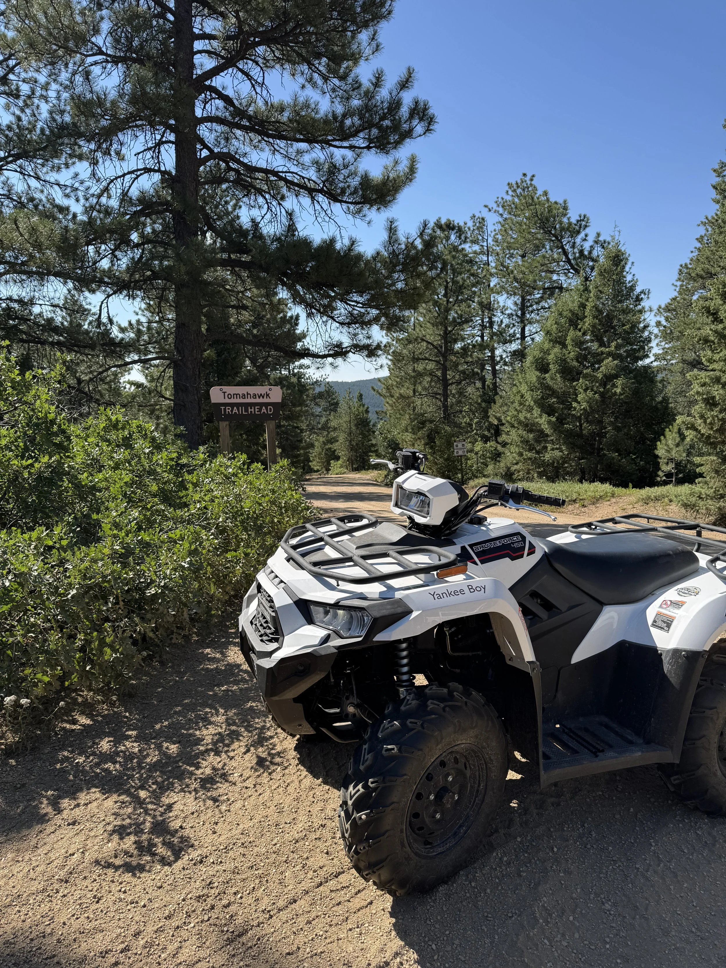 An ATV parked on a dirt trail in a forested area with tall pine trees, a blue sky, and a sign that reads 'Tomahawk Trailhead' in the background.