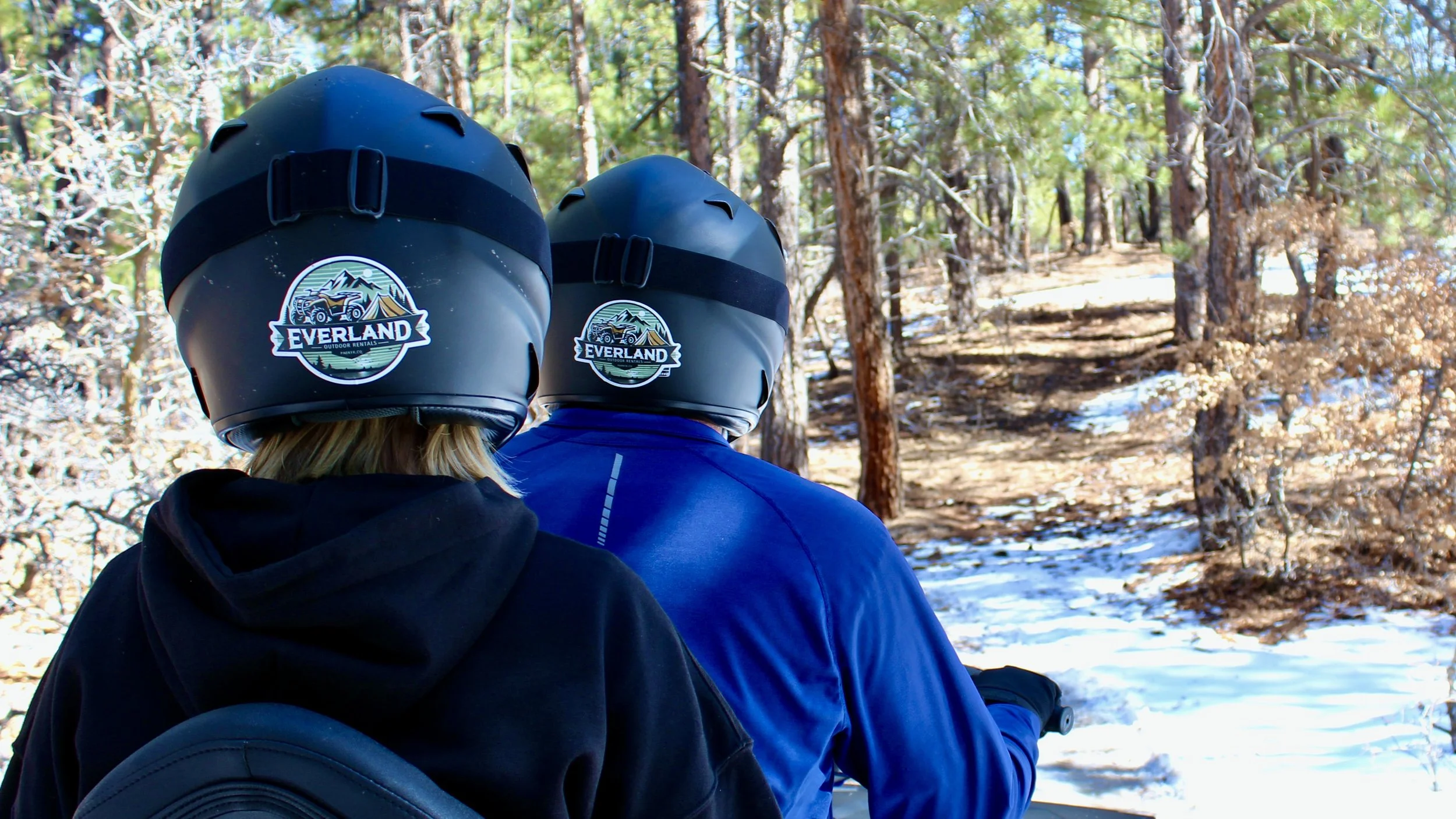 Two people wearing black helmets with the Everland logo, riding an ATV through a forest trail with trees and patches of snow.