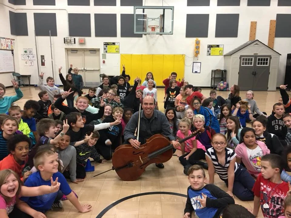 A group of children sitting and lying on the gymnasium floor, smiling and making peace signs, around a man playing a cello.